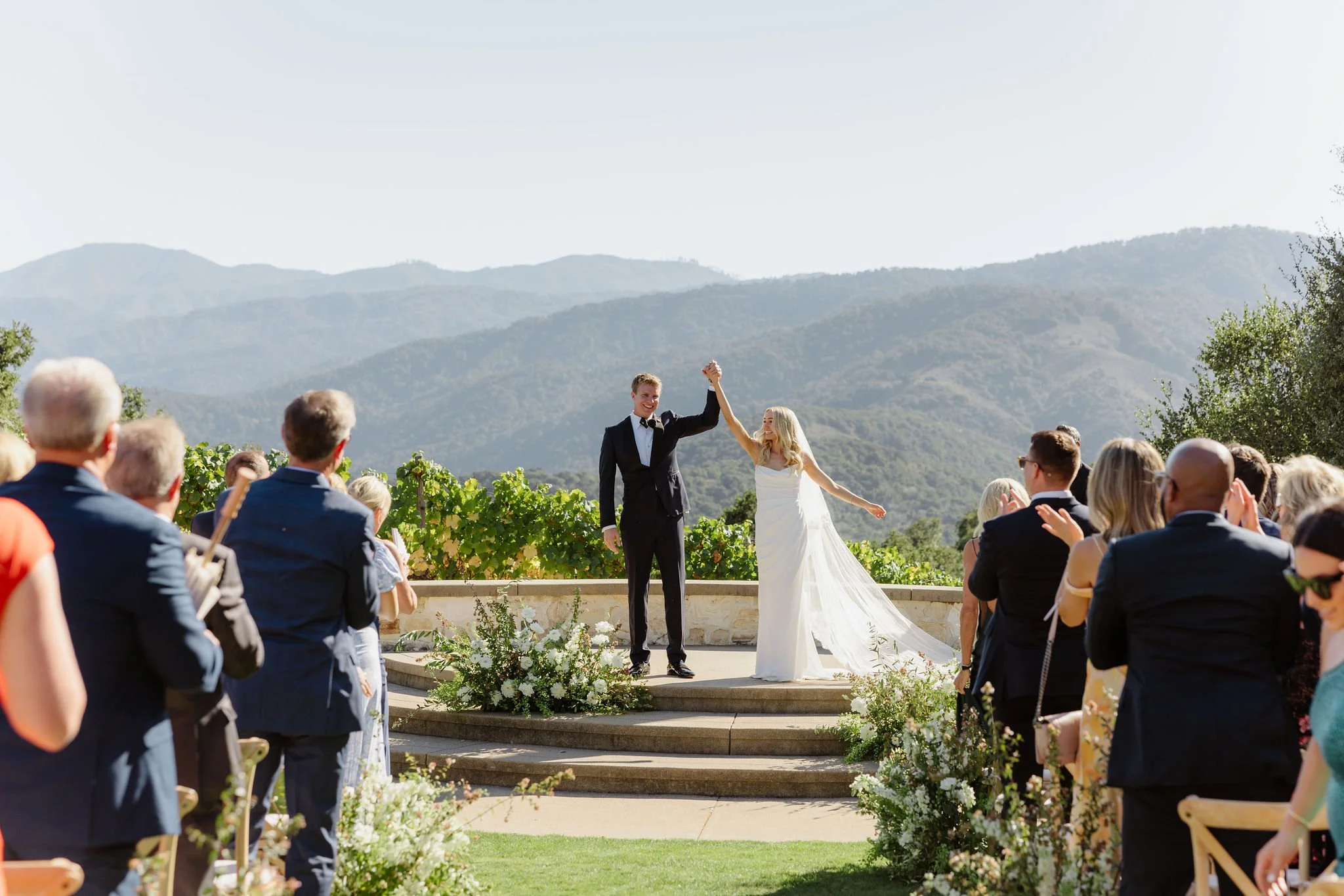 A couple, a groom and a bride, holding hands and smiling at each other while standing on a small stage during a wedding ceremony outdoors, surrounded by guests with scenic mountains in the background.