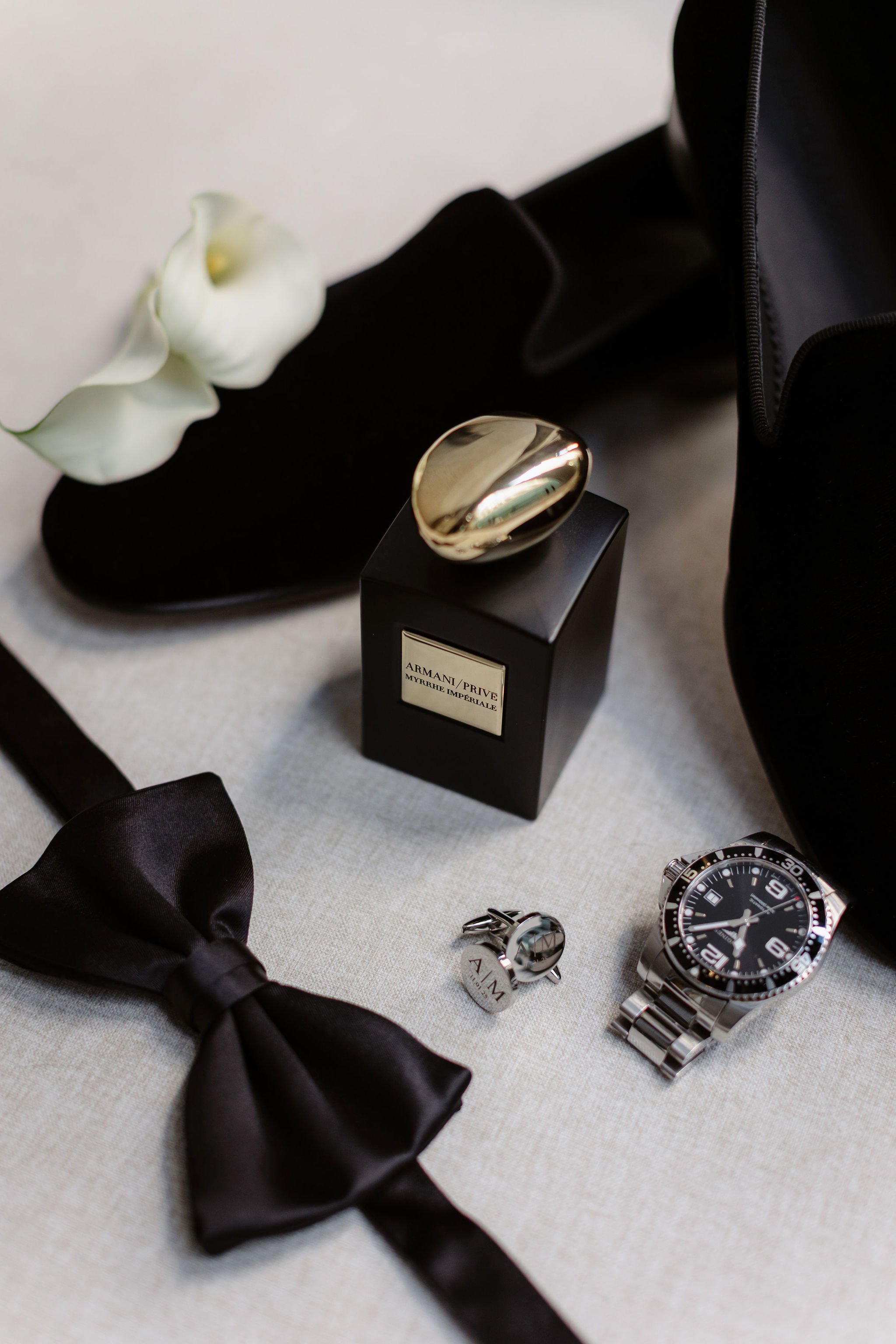 A black satin bowtie, a silver wristwatch with a black face, a pair of cufflinks, a perfume bottle labeled Armani Privé Myrrhe Imperiale, a white calla lily flower, and a black garment or accessory on a light-colored table.