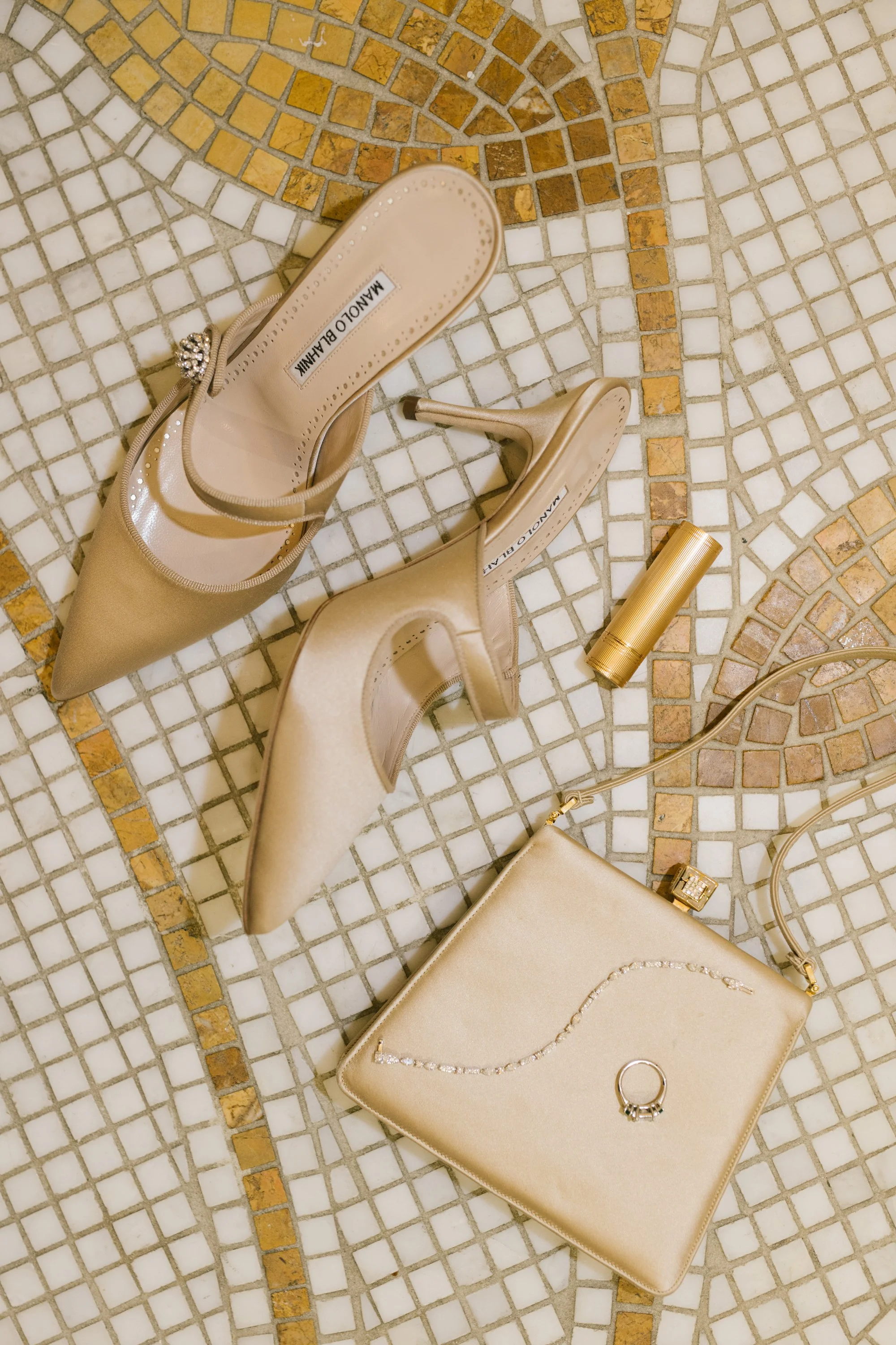 A pair of beige high-heeled shoes, a gold lipstick tube, a beige purse with a gold chain, and some jewelry are arranged on a mosaic tiled floor.