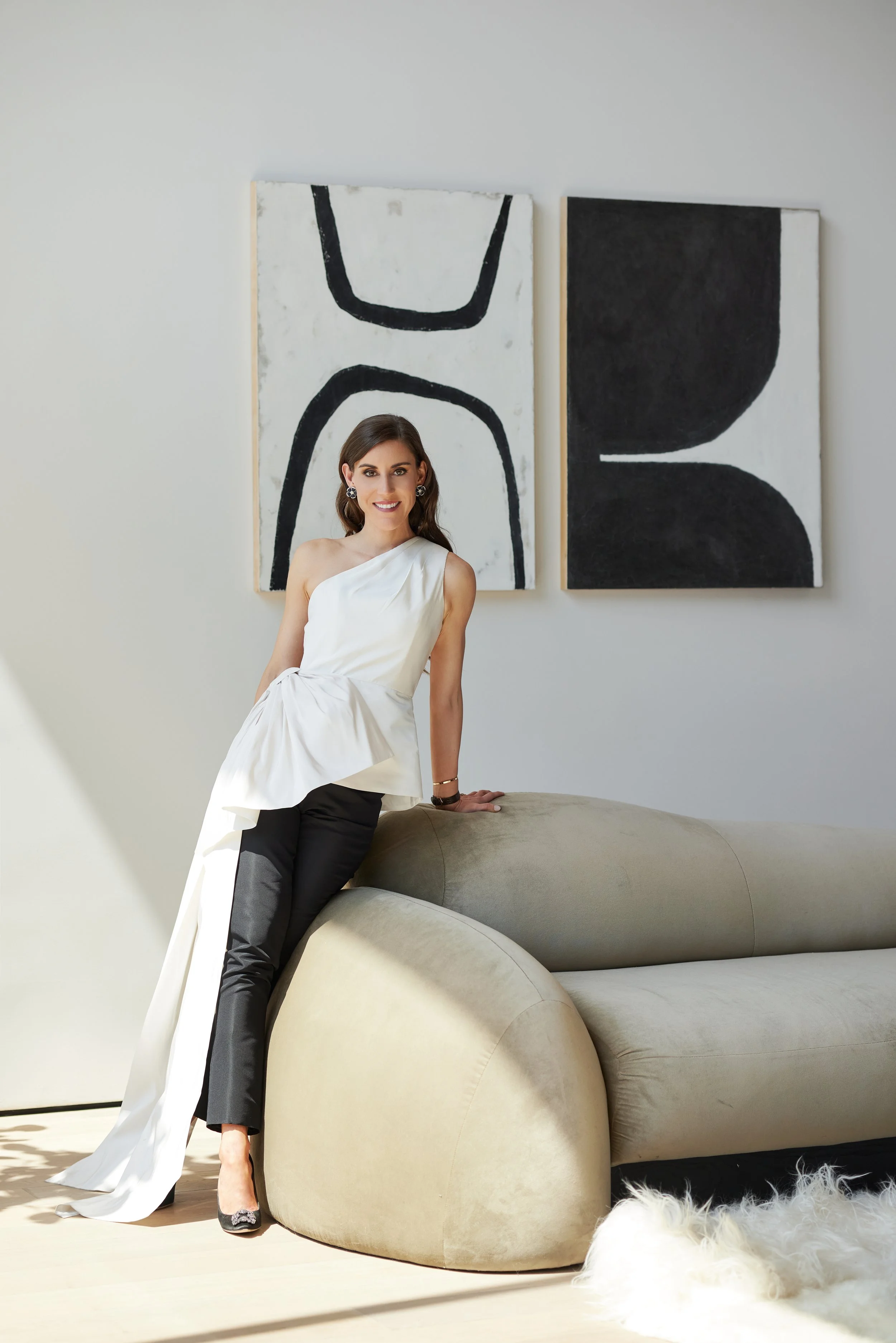 A woman in a white one-shoulder top and black pants sitting on a beige curved sofa in a modern, minimalist room with abstract black and white paintings on the wall behind her, and a furry white dog lying on the light-colored wooden floor nearby.