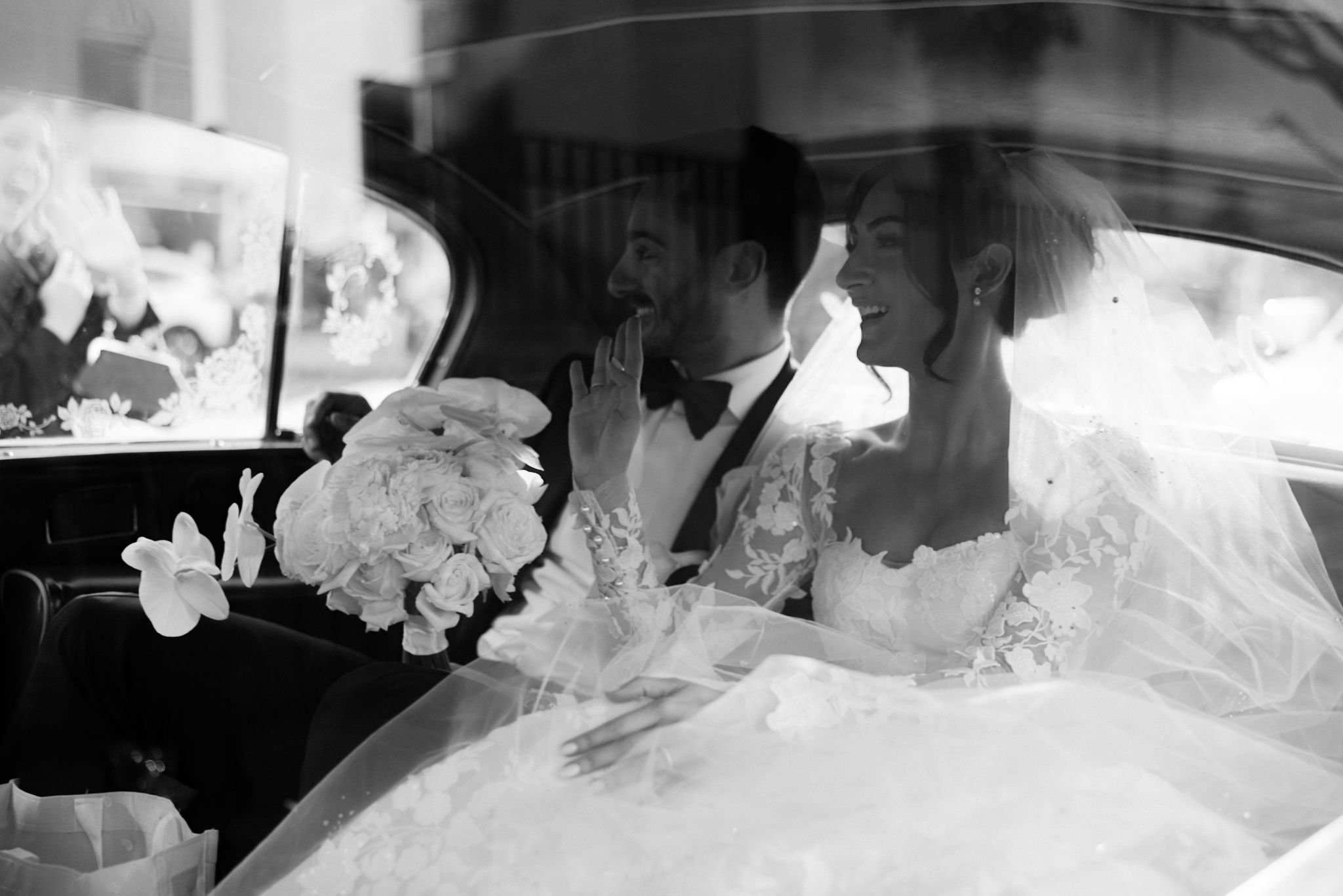 A black and white photo of a bride and groom sitting in the back of a car, smiling at each other. The bride is holding a bouquet of flowers. They are dressed in wedding attire. The window shows reflections and nearby objects.