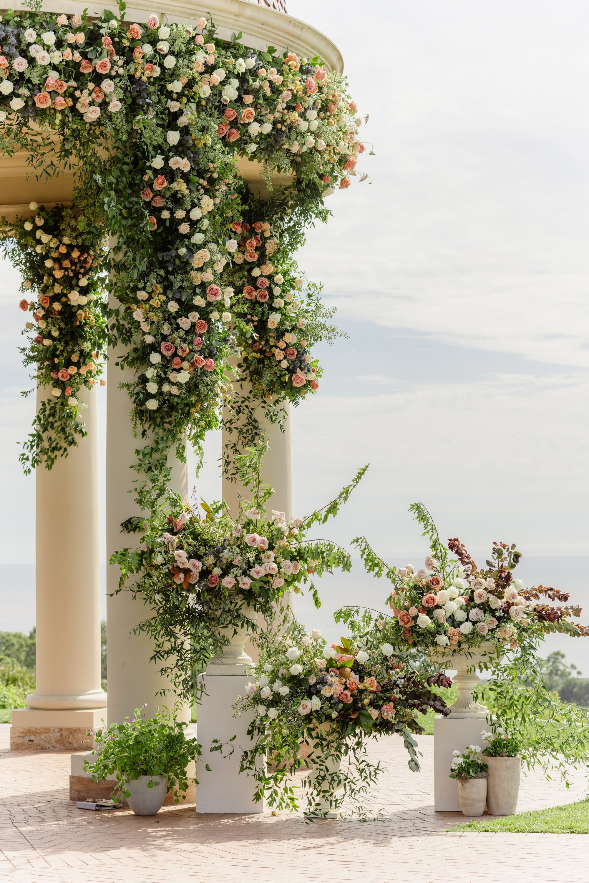 Floral arrangements on white columns for a wedding or event, with greenery and pink, white, and purple flowers, set outdoors on a brick patio.