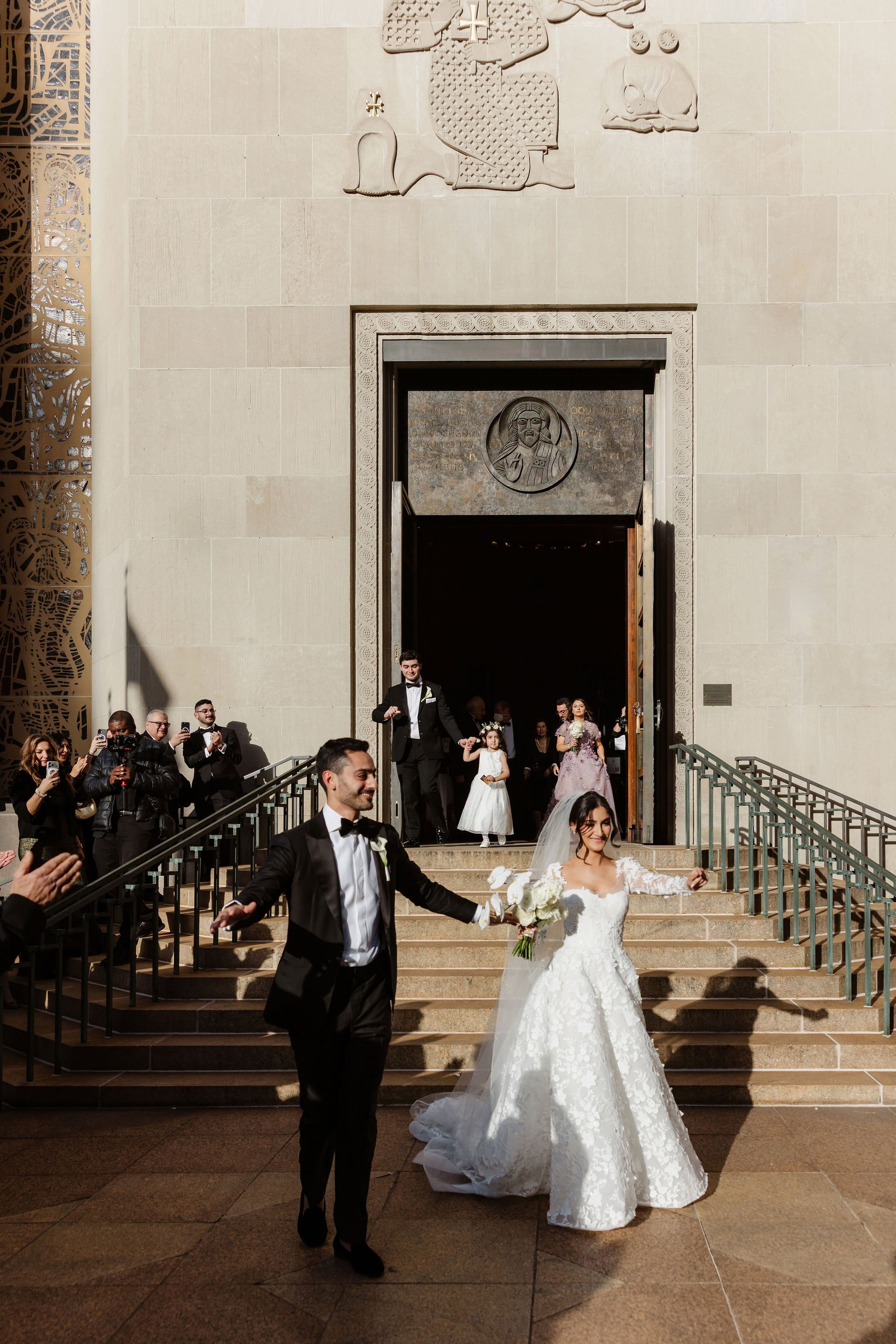 A bride and groom exiting a church or cathedral after their wedding ceremony, with a group of guests and photographers taking pictures and celebrating on the stairs outside.