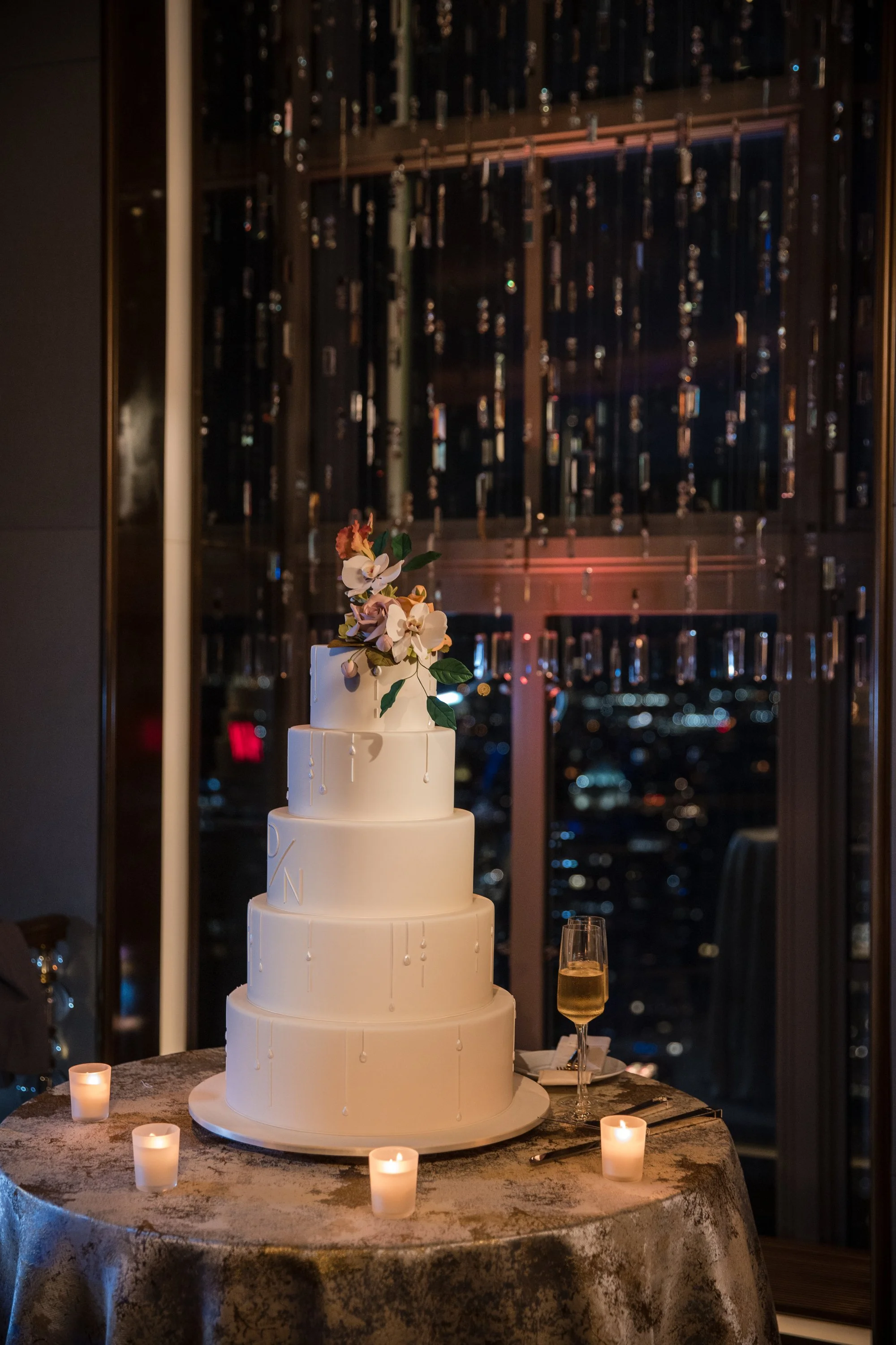 A multi-tiered white wedding cake with floral decorations on top, surrounded by small lit candles on a table with a metallic tablecloth, overlooking a city view at night through large windows.