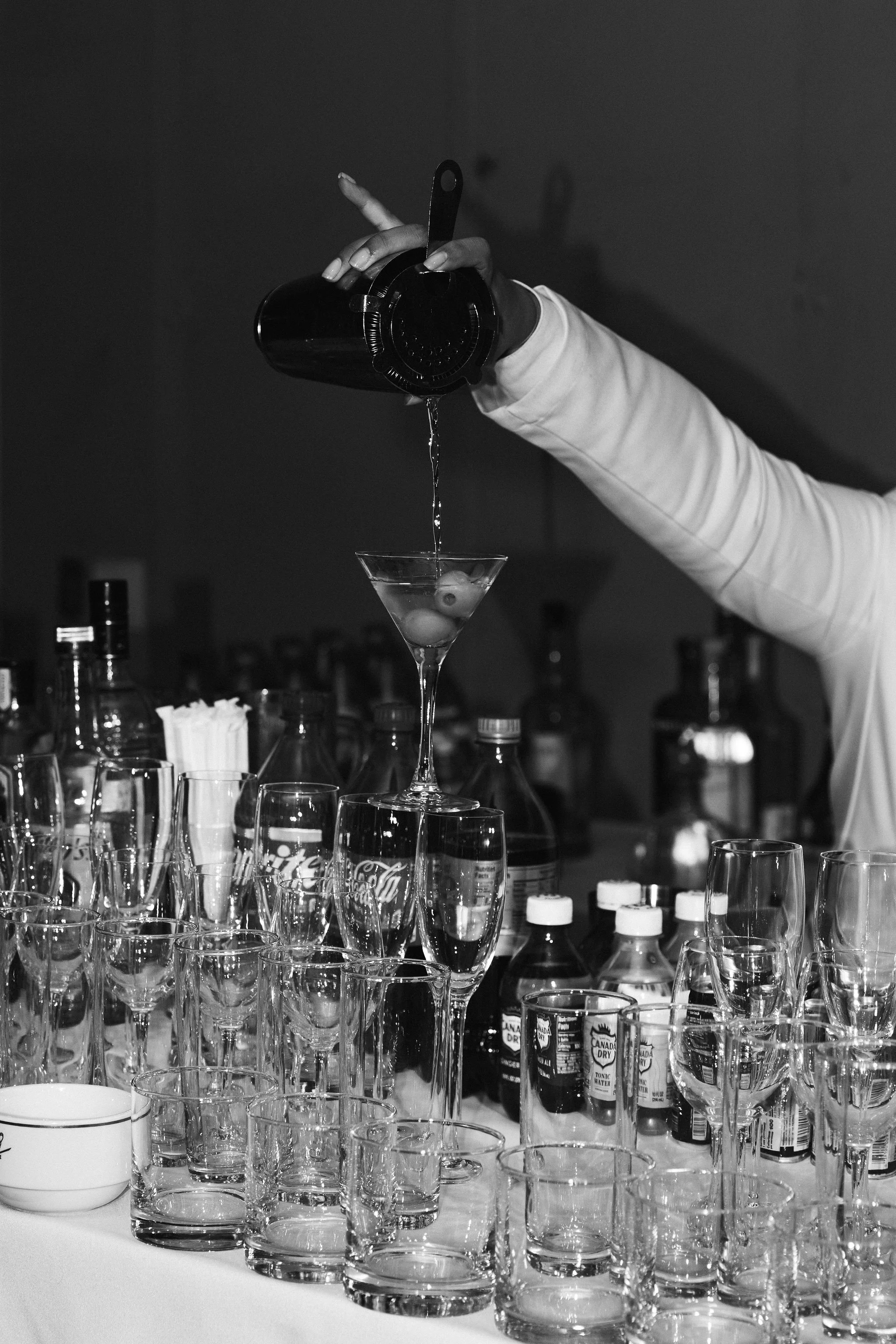A hand in a coat sleeve pouring a liquid from a cocktail shaker into a martini glass, with several glasses and bottles on the table.