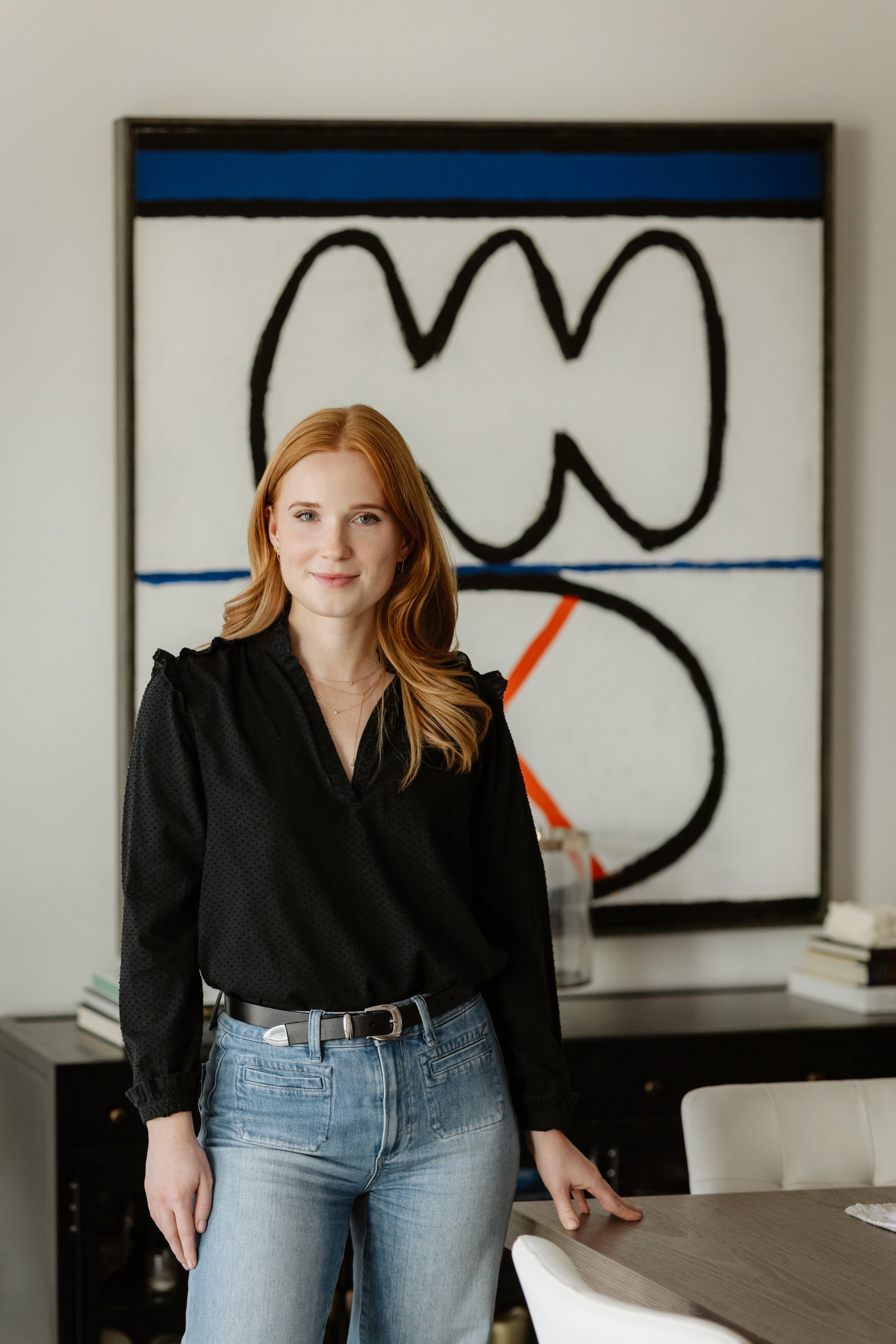 A young woman with red hair and fair skin is standing in a modern office or conference room. She is wearing a black blouse with puffed sleeves and high-waisted blue jeans with a black belt. Behind her is an abstract painting with black, white, blue, 