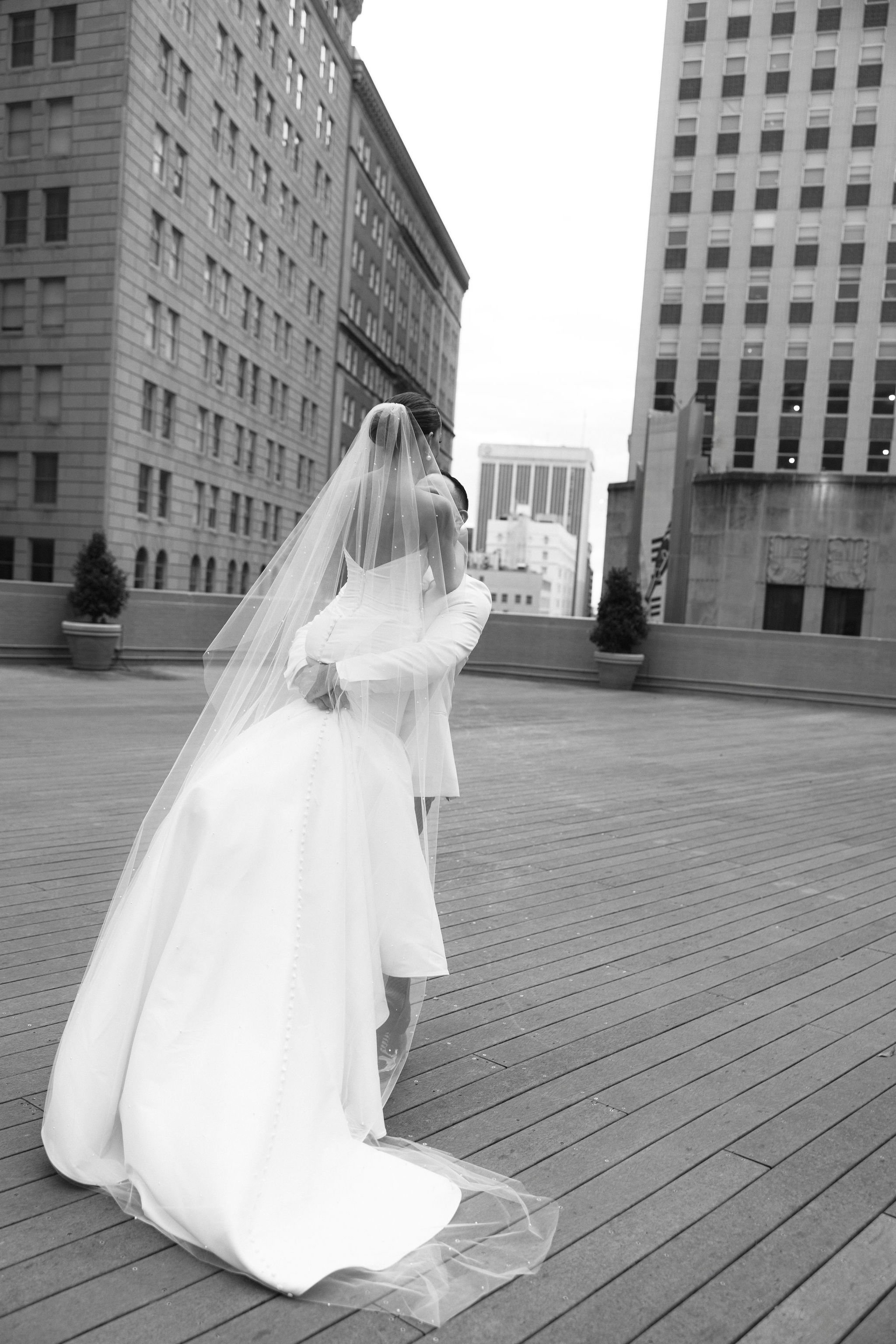 A bride in a wedding dress and veil embraces on a rooftop with city buildings in the background.