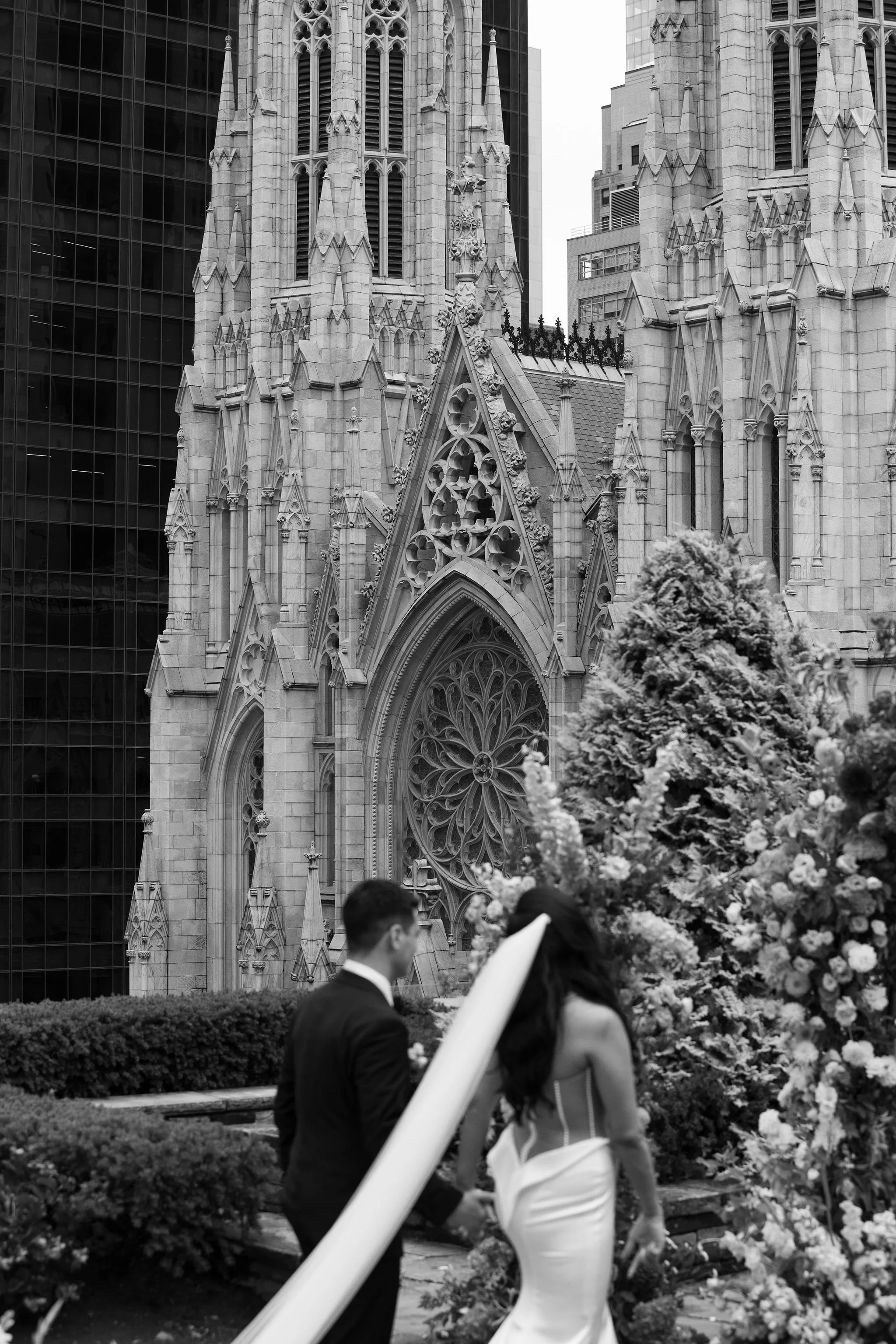 A black and white photo of a Gothic-style church with a tall steeple, surrounded by modern glass buildings. In the foreground, a couple is walking past a floral display.