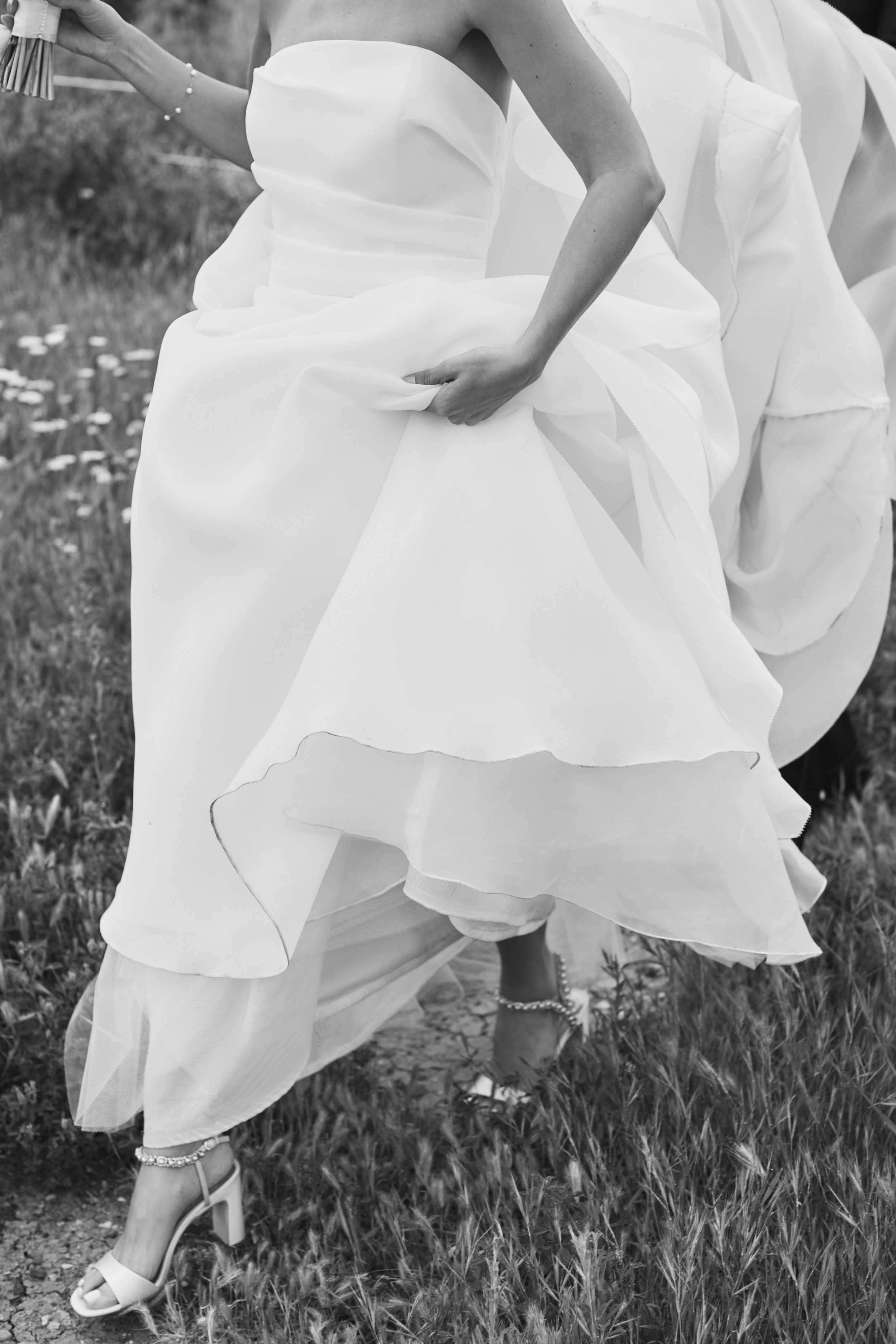 A person wearing a strapless white dress with layers and a full skirt, holding the skirt up slightly with one hand while walking outdoors on a grassy surface, wearing high-heeled shoes and jewelry, in black and white.