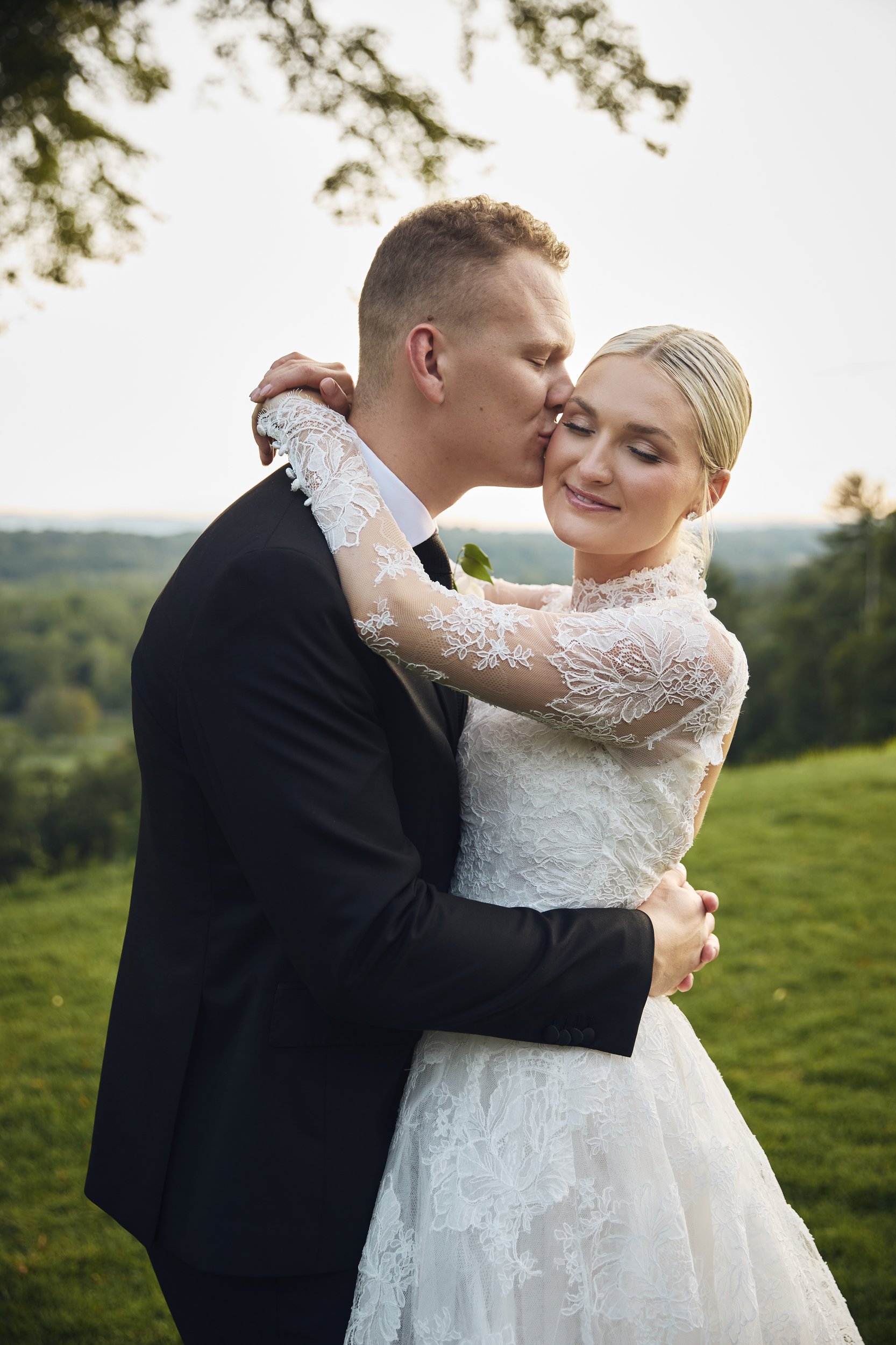 A newlywed couple sharing a kiss outdoors on a grassy hill with trees and a sunset sky in the background. The groom is wearing a black suit and the bride is in a white lace wedding dress with long sleeves.