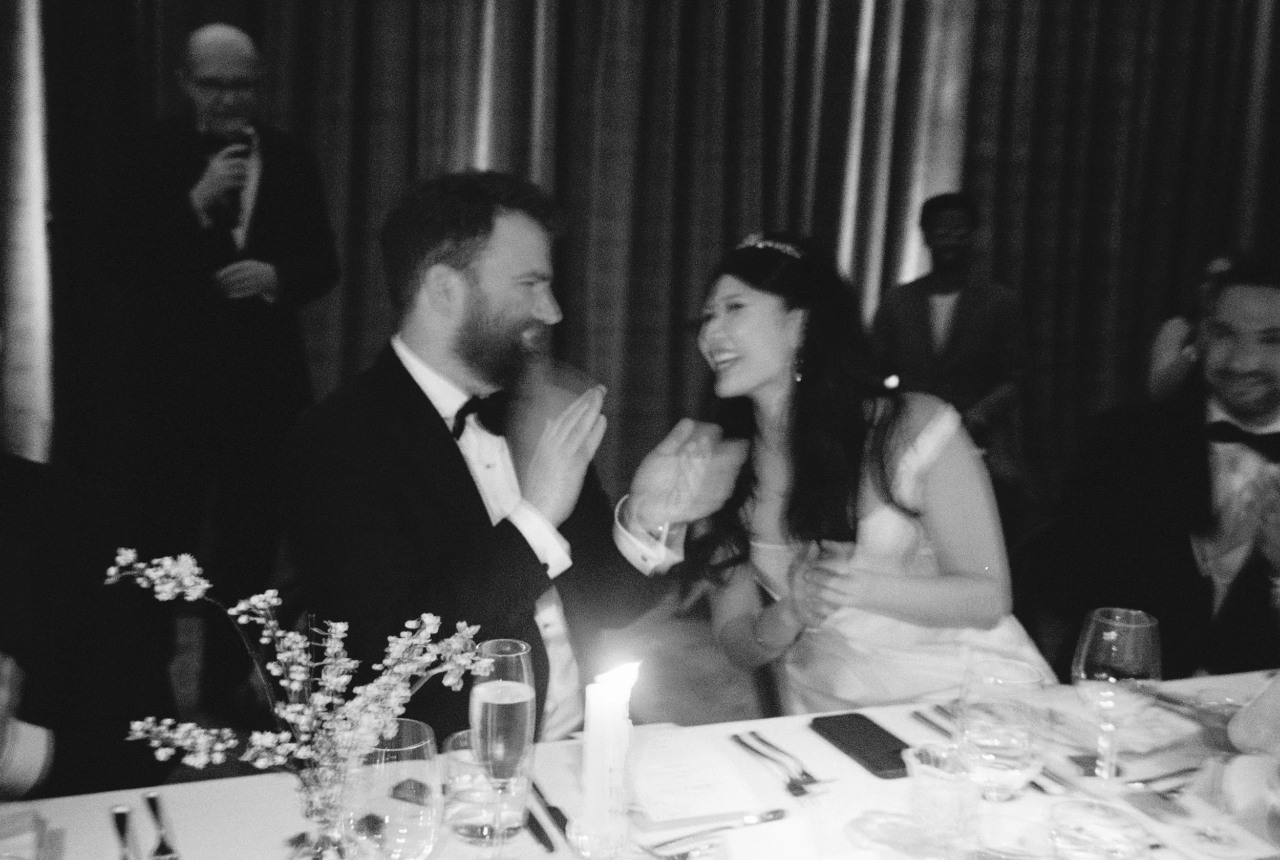 A black and white photo of a wedding reception with a bride and groom smiling and talking at the table, surrounded by guests and floral decorations.