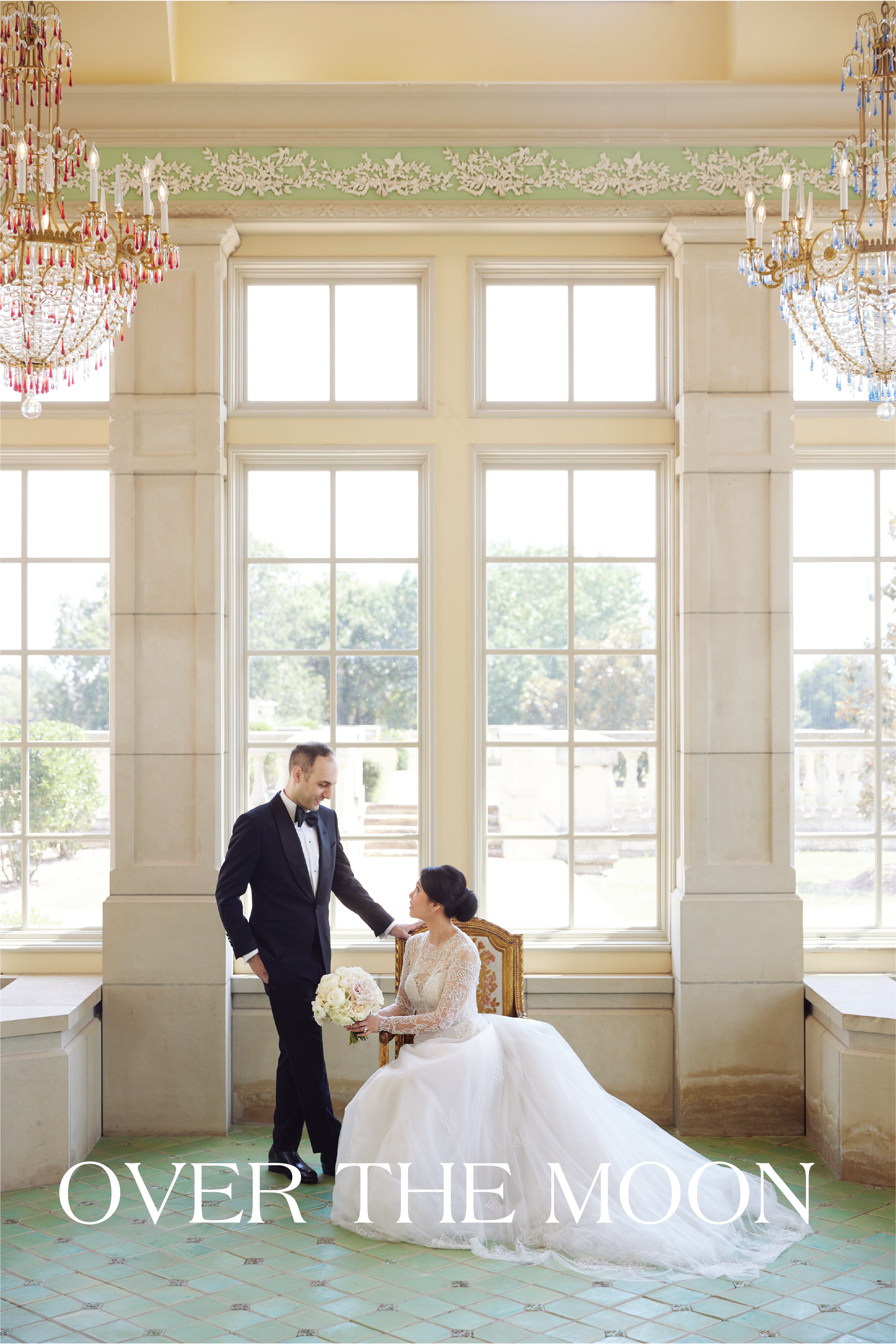 A bride and groom in a bright, elegant room with large windows and chandeliers. The groom stands in a tuxedo, holding the bride's hand, who is seated in a white wedding gown holding a bouquet of white roses. The scene appears romantic and formal, likely part of a wedding ceremony or photo shoot.