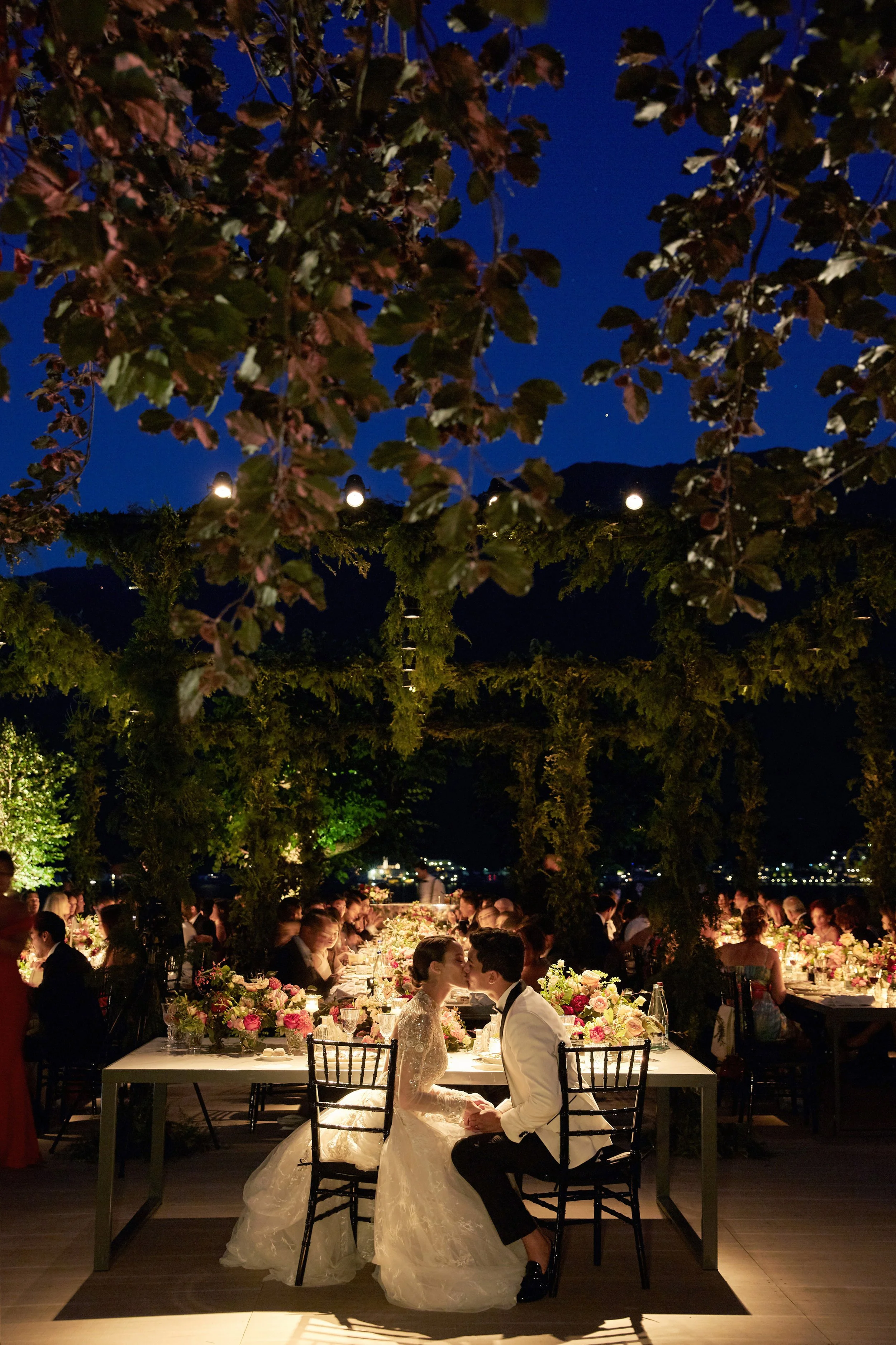 A bride and groom sit side by side in an outdoor wedding reception at night, holding hands and gazing at each other, with a beautifully decorated event under string lights and trees, surrounded by many guests.