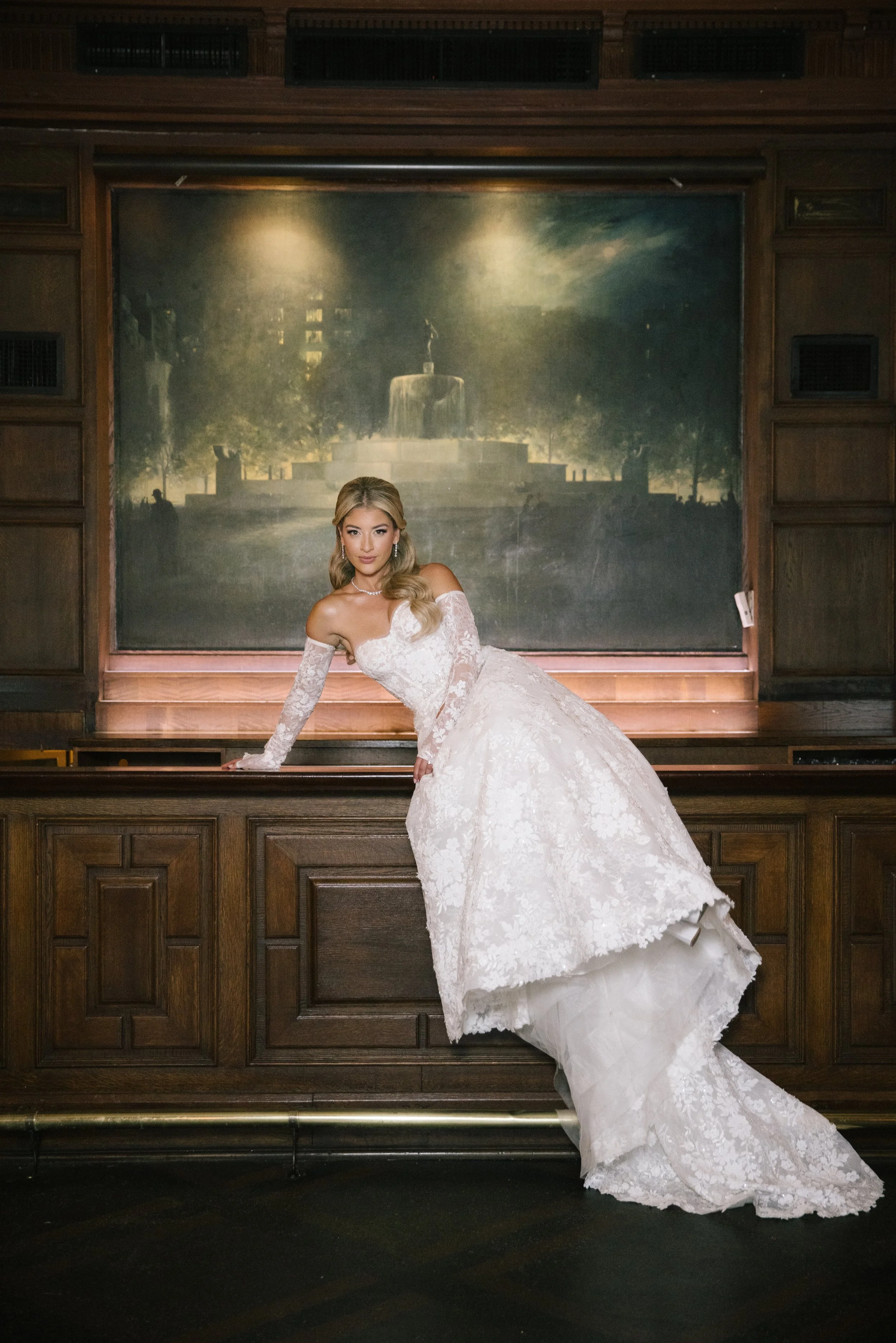 A woman in a white lace wedding dress posed in front of a large, historic painting of a fountain in a wooden frame.