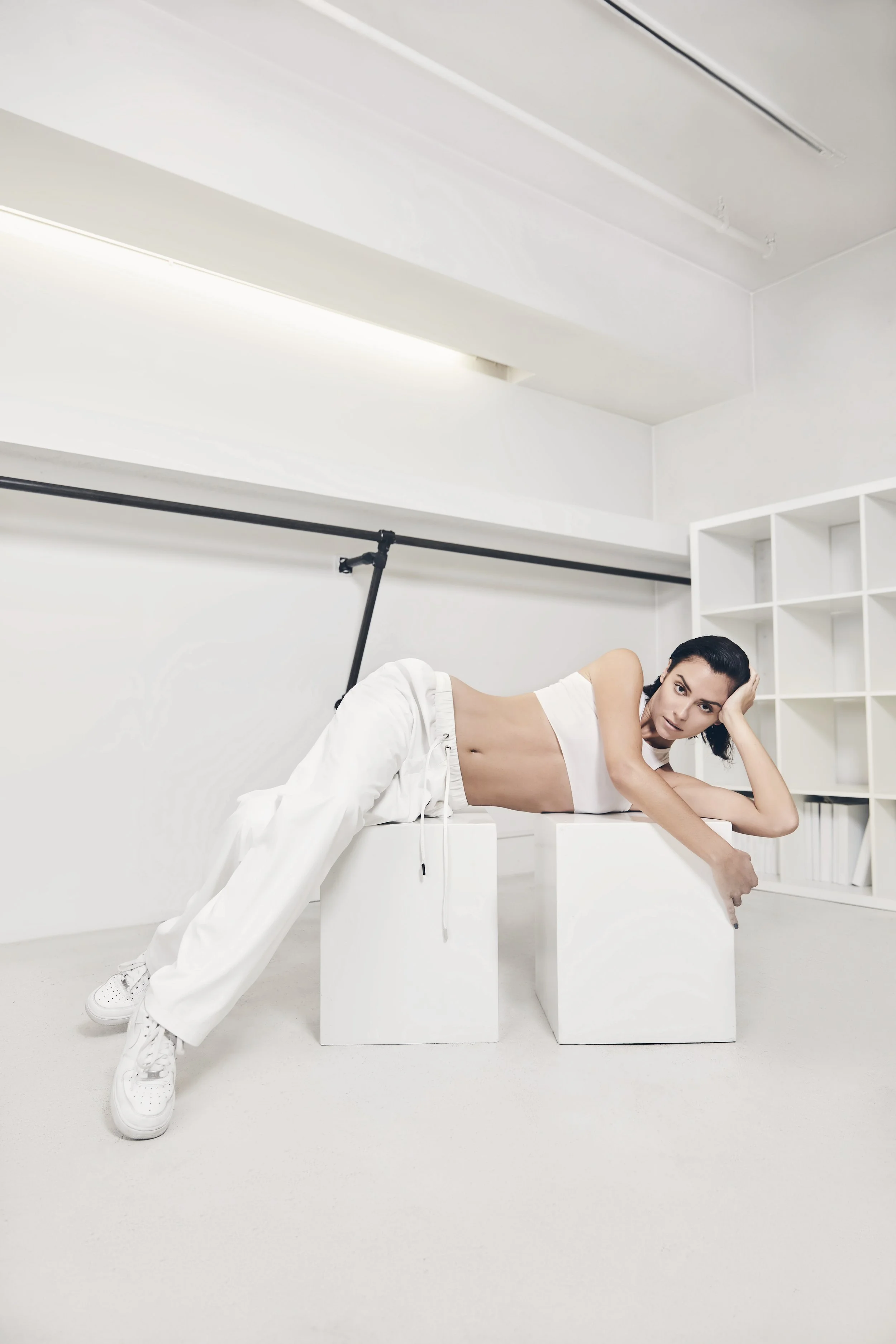 A woman in white cropped top, white pants, and sneakers lies on white blocks in a white minimalist room, propped on one elbow with her hand on her head, looking at the camera.