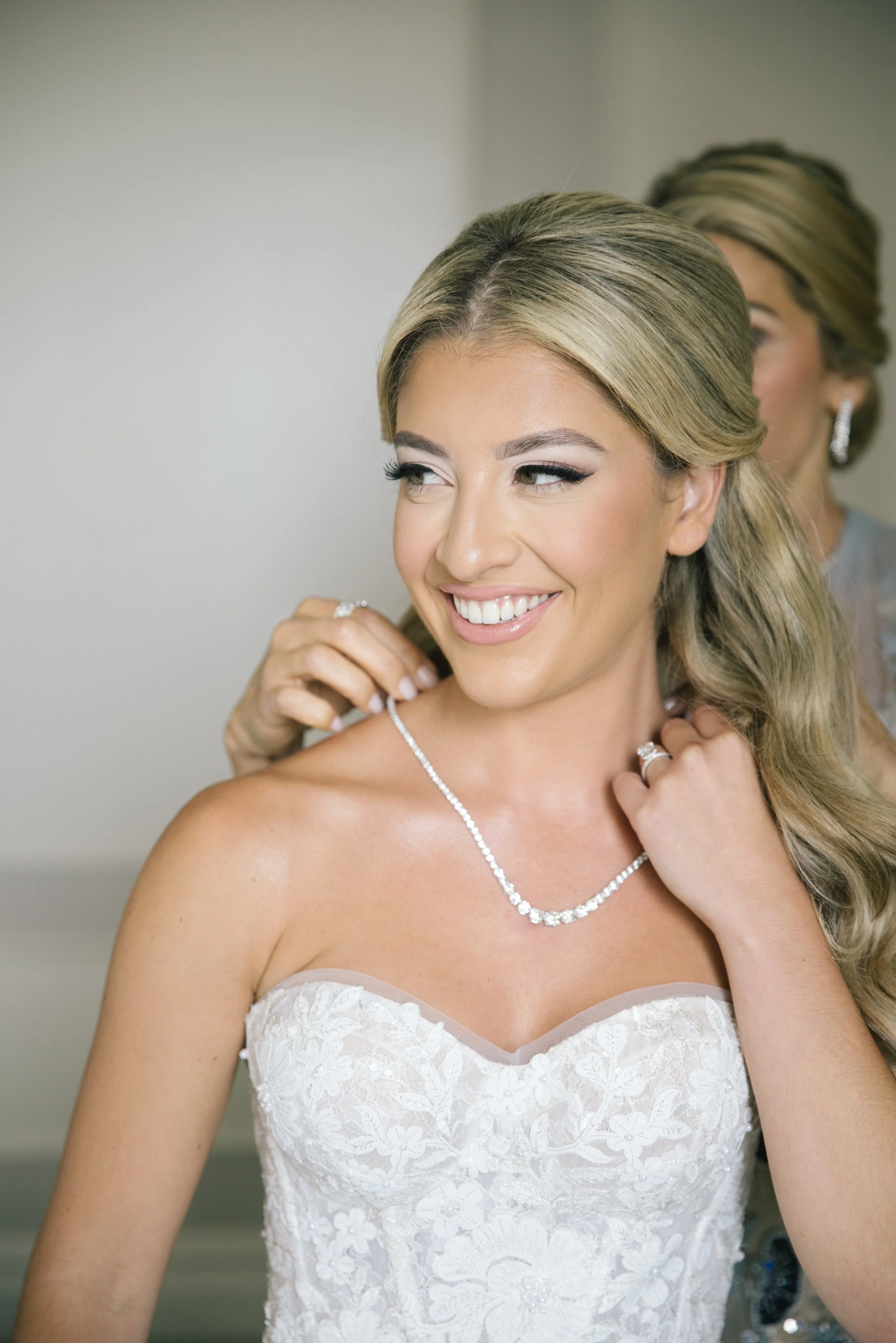 A bride with blonde hair smiling, wearing a white lace wedding dress and jewelry, with another woman in the background preparing her for the wedding.