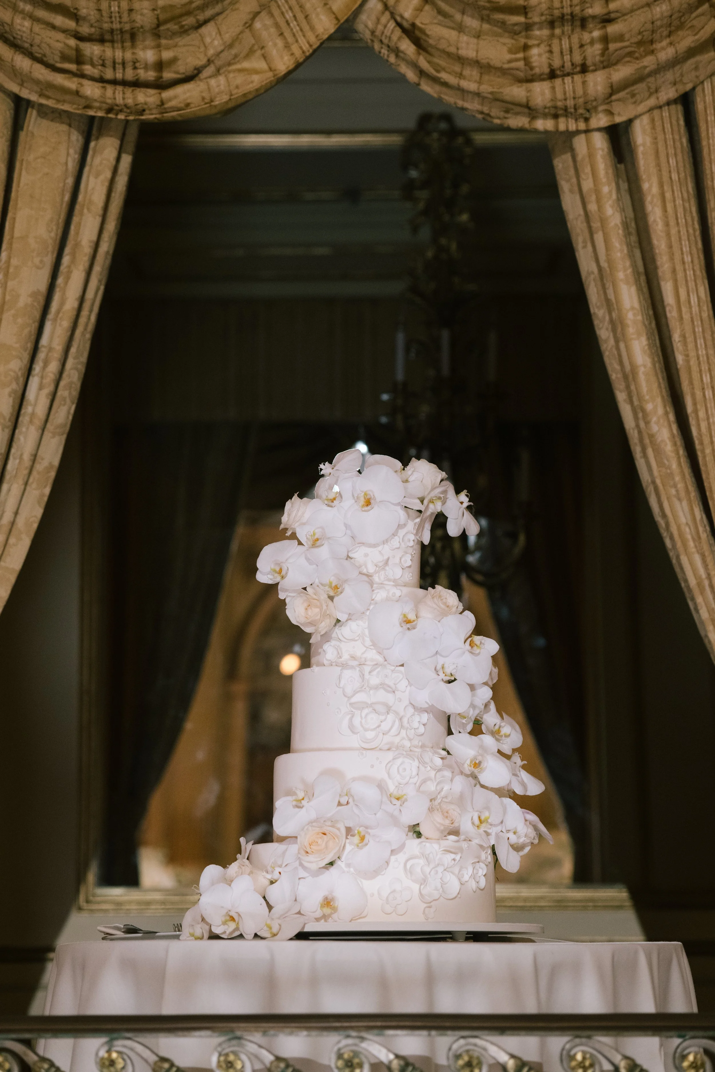 A white floral wedding cake with orchids on top, multiple tiers, on a table with a white tablecloth, set against an ornate mirror and draped curtains.