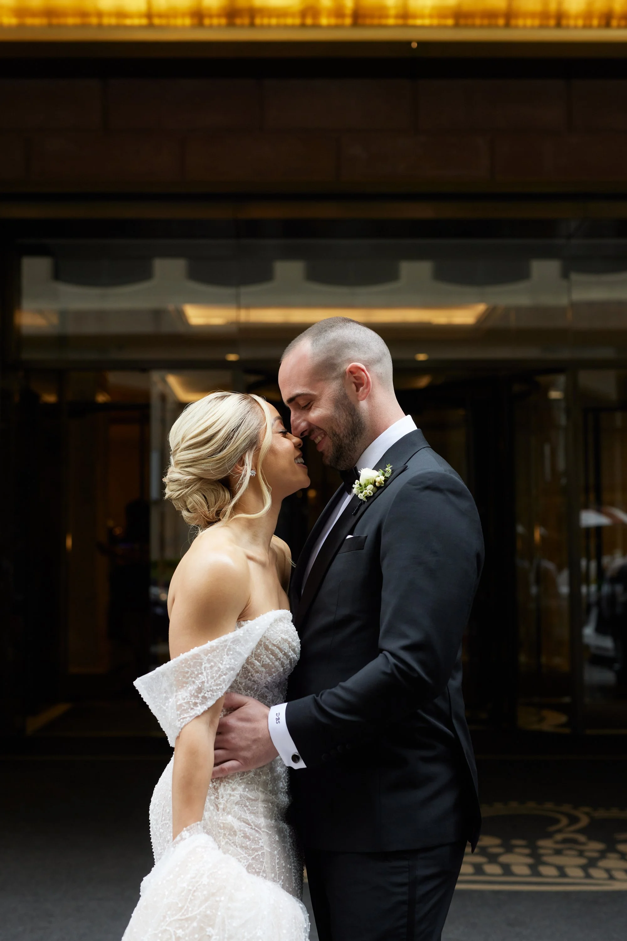 A bride and groom are embracing and smiling at each other in an indoor setting, with the groom dressed in a black tuxedo and the bride in a strapless lace wedding gown.