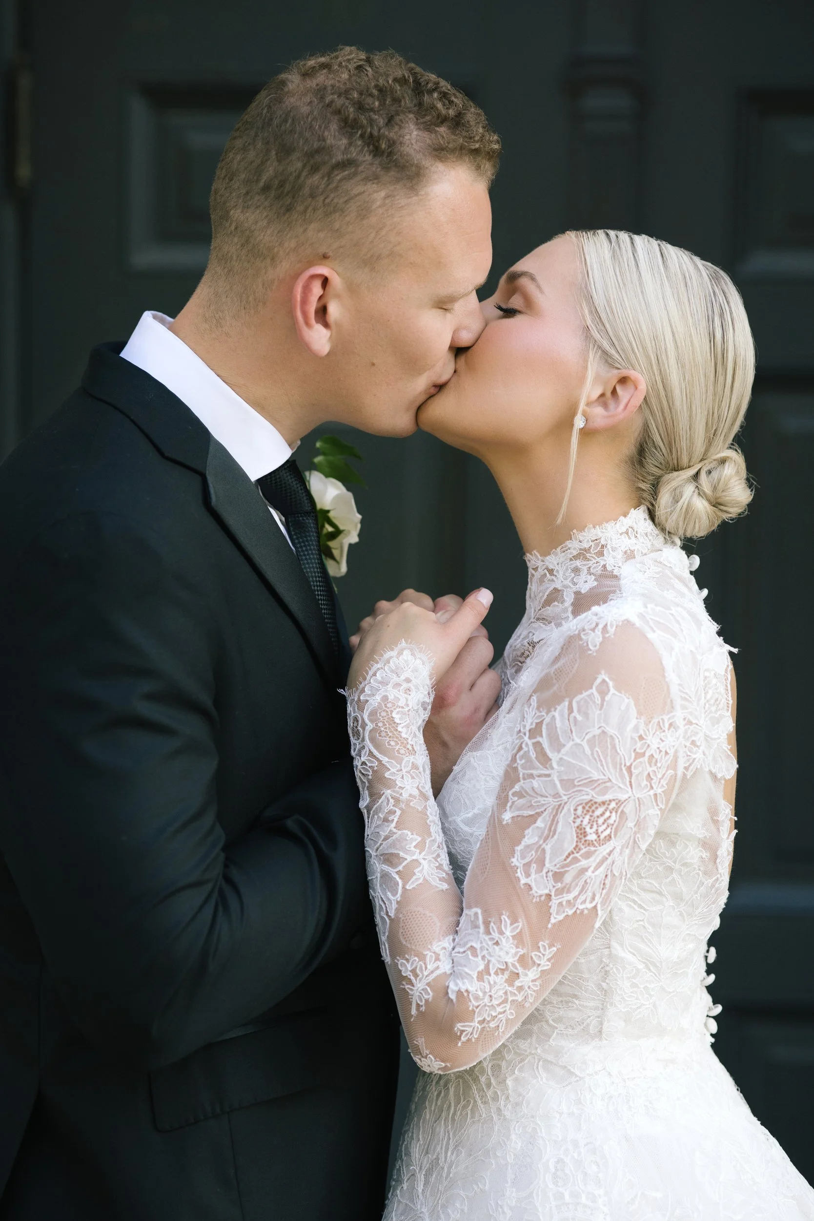 A bride and groom share a kiss during their wedding, with the bride wearing a lace wedding dress and the groom in a black suit and tie.