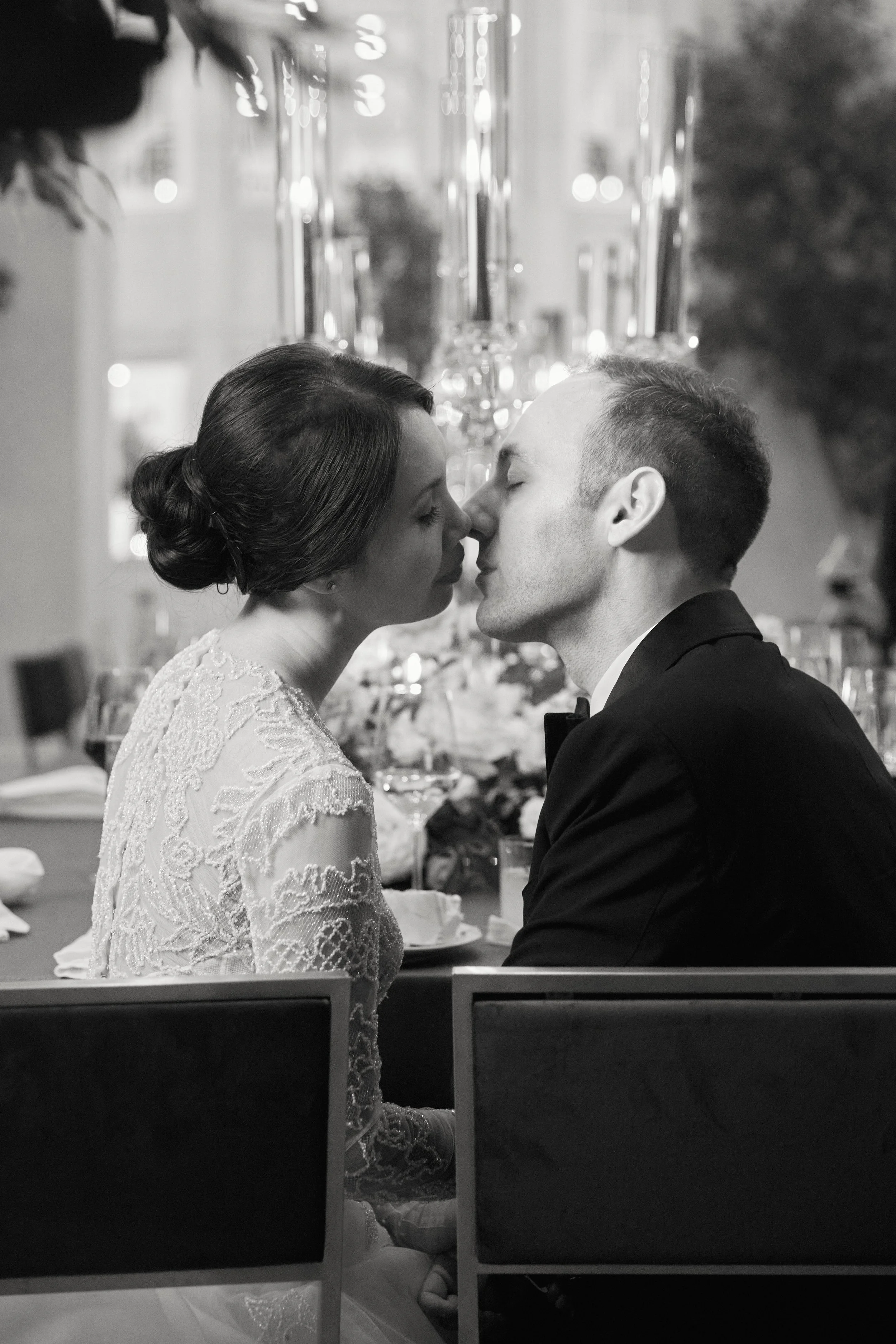 Two people, a woman in a lace wedding dress and a man in a tuxedo, are kissing at a wedding reception. The background features candlelit decorations and blurred lights.