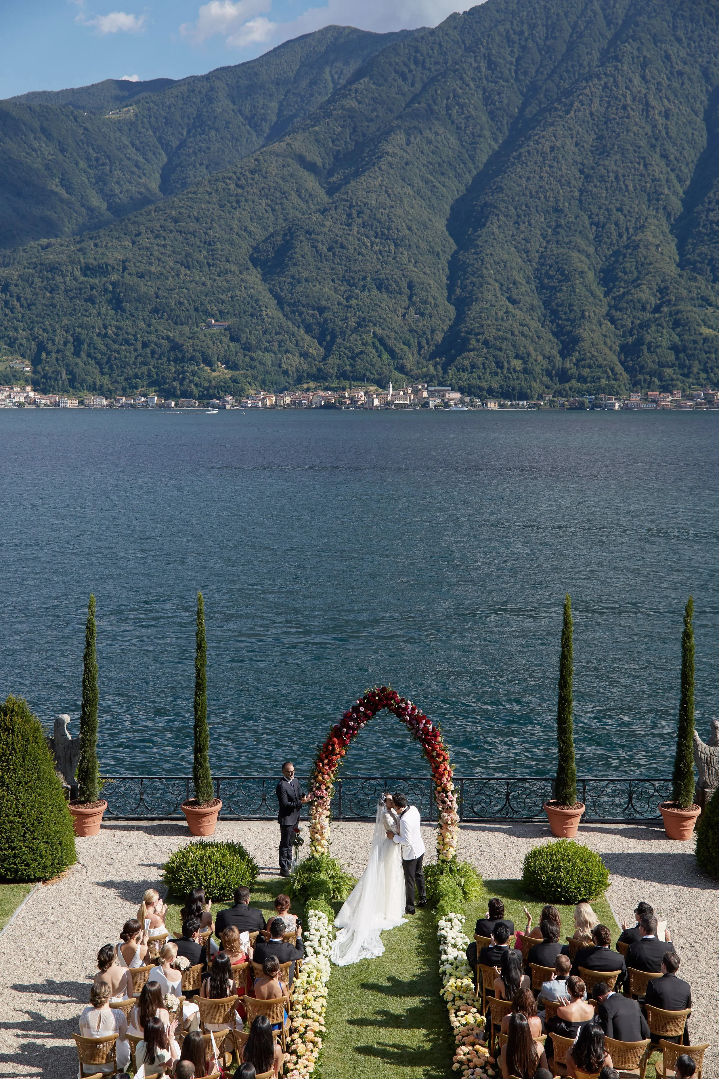 A wedding ceremony taking place outdoors by a lake with mountains in the background. The bride and groom stand under a floral arch, surrounded by guests seated in chairs on a grassy lawn. Twelve tall potted plants embellish the scene, with a gravel p