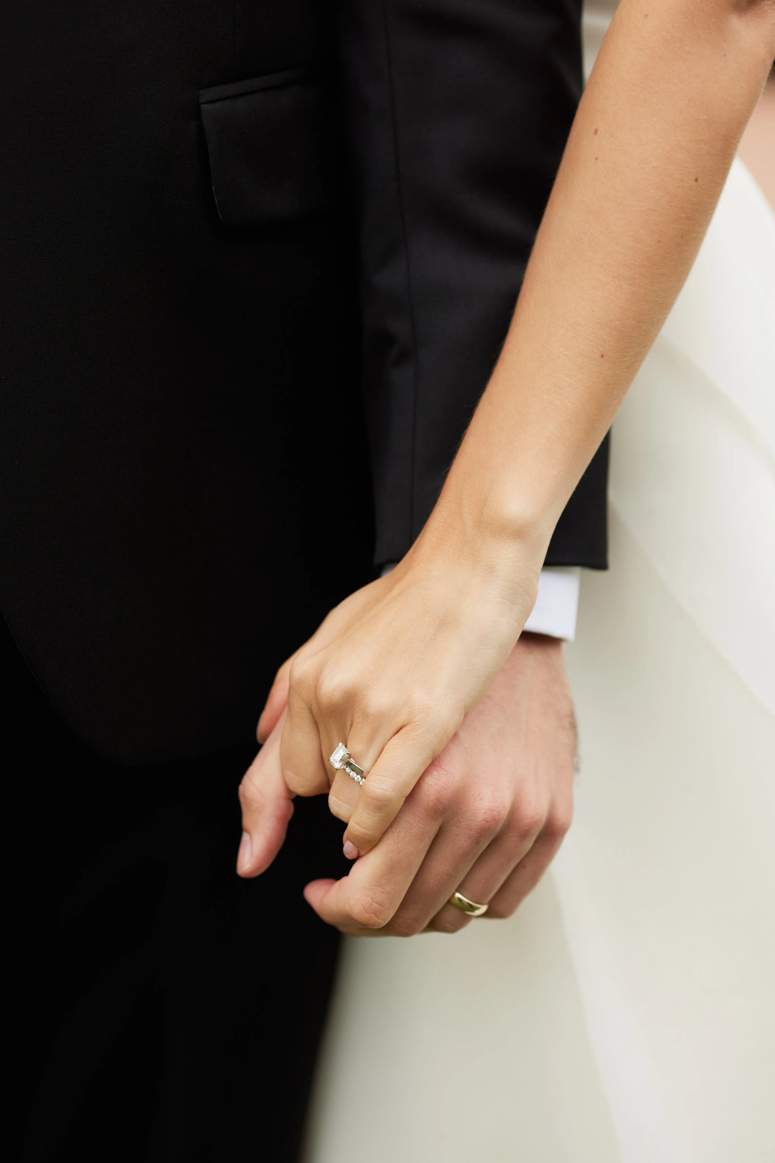 A close-up of a couple holding hands. The woman's hand shows a large, shimmering engagement ring with a prominent gemstone. The man is wearing a plain band on his ring finger.
