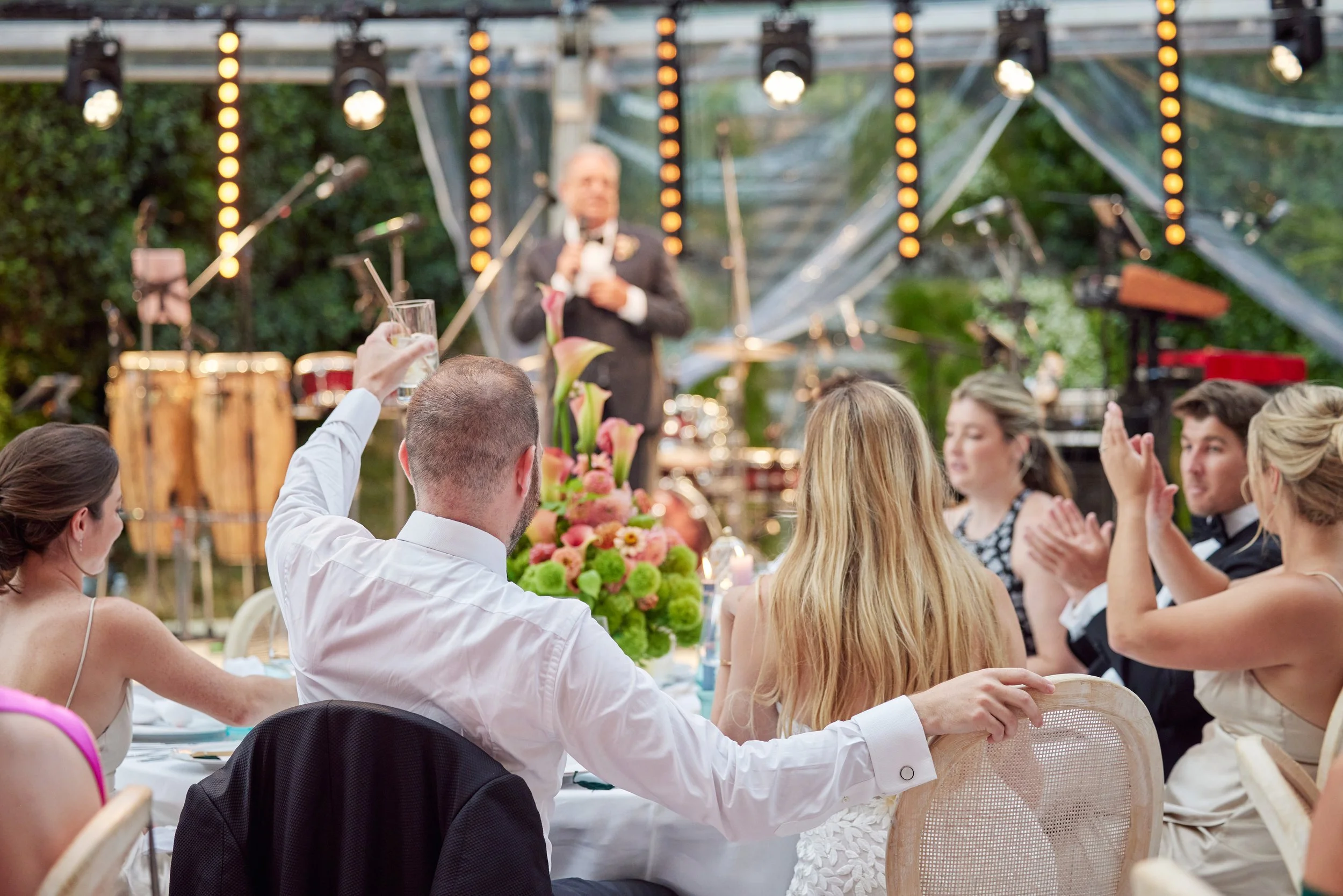 Guests seated at a long table during an outdoor celebration, with a guitarist and a speaker in the background on a decorated stage surrounded by greenery.