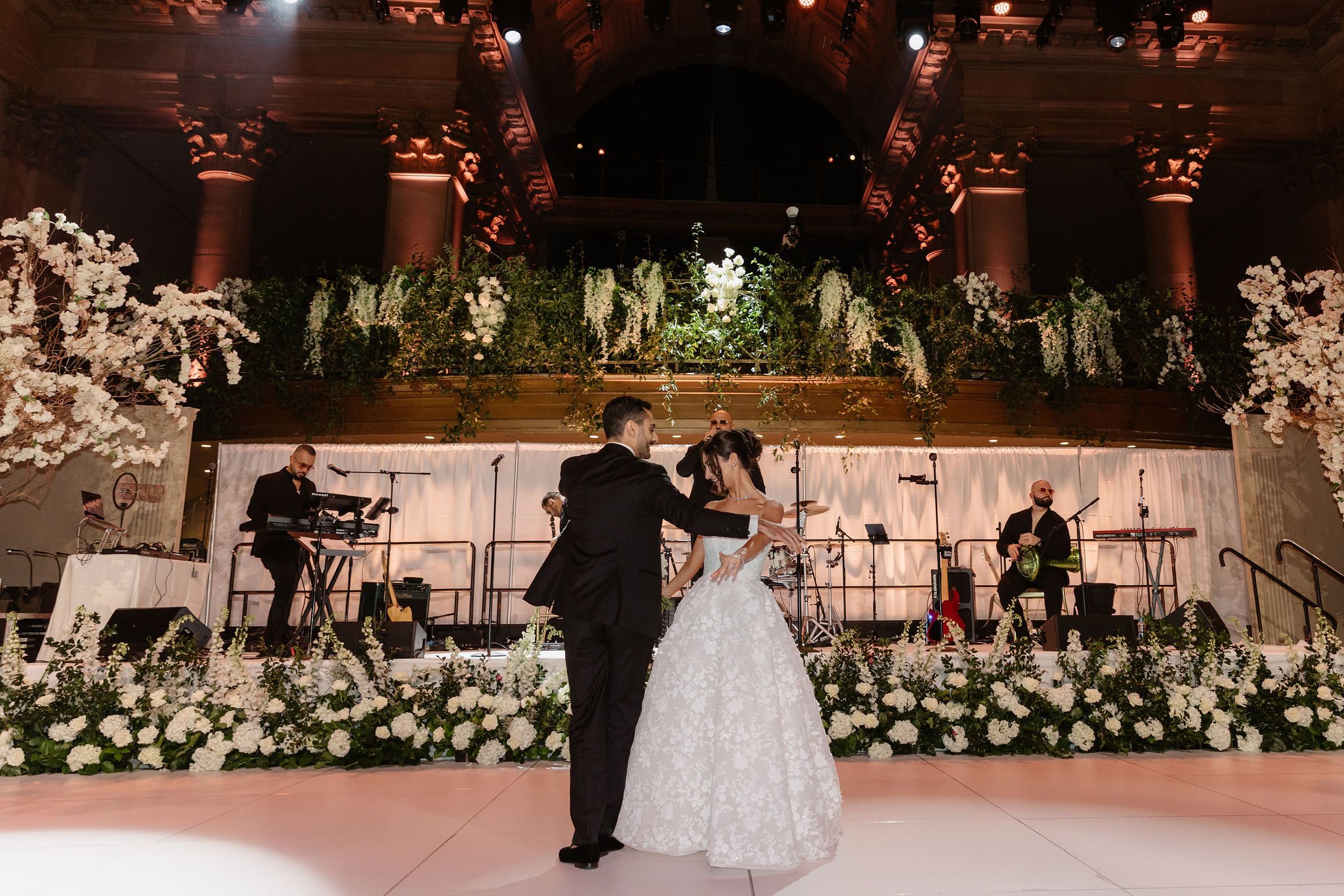 A bride and groom are dancing together on a stage decorated with white flowers. A band of musicians is playing in the background on a stage with a floral and green foliage backdrop, in a grand hall with ornate architecture and lighting.
