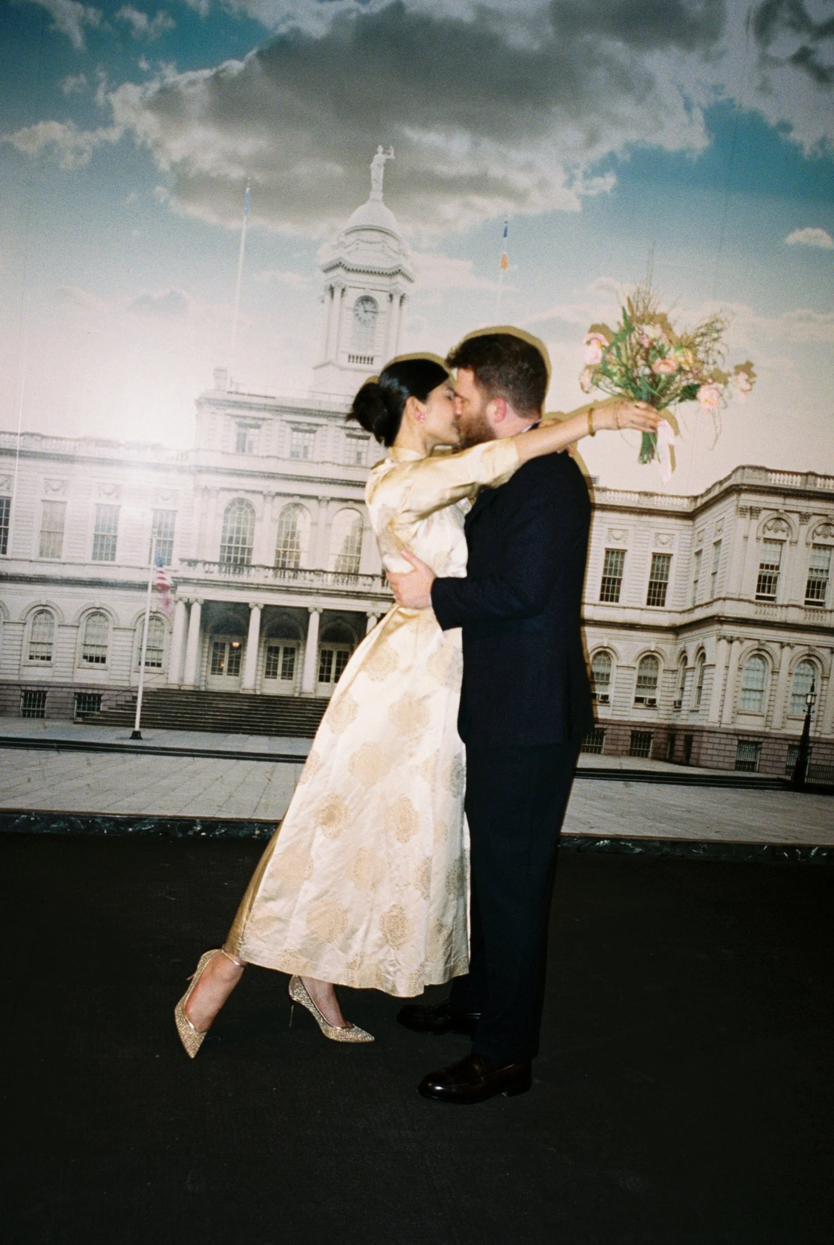 A couple is dancing closely in front of a backdrop featuring a historic government building with a clock tower, flags, and a cloudy sky. The woman is wearing a gold-colored evening gown with high heels, and the man is dressed in a dark suit.