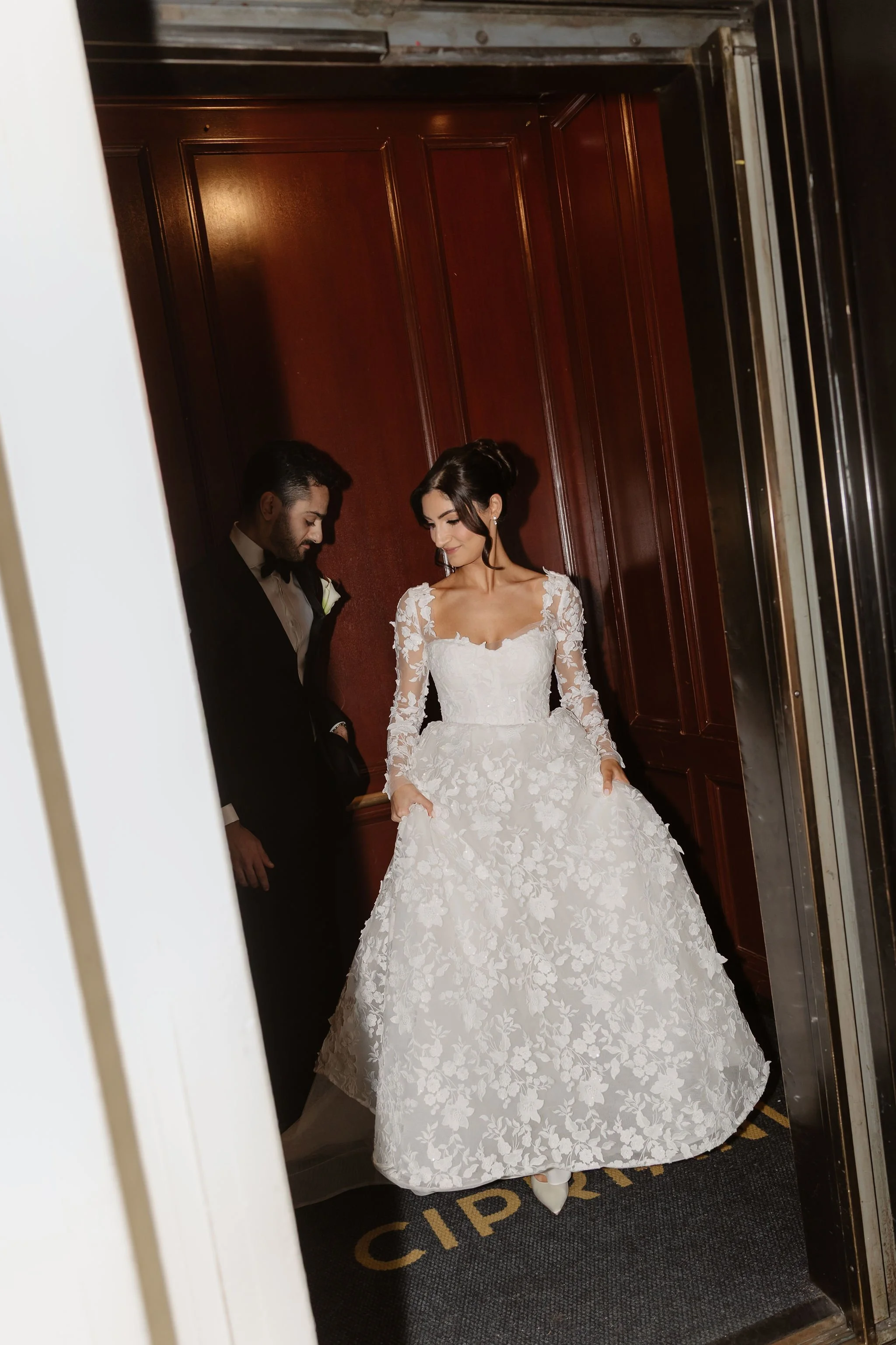 A bride in a white lace wedding gown with long sleeves and floral patterns, holding the sides of her dress, and a groom in a black tuxedo with a bow tie, standing close together in an elevator with wooden panel walls.