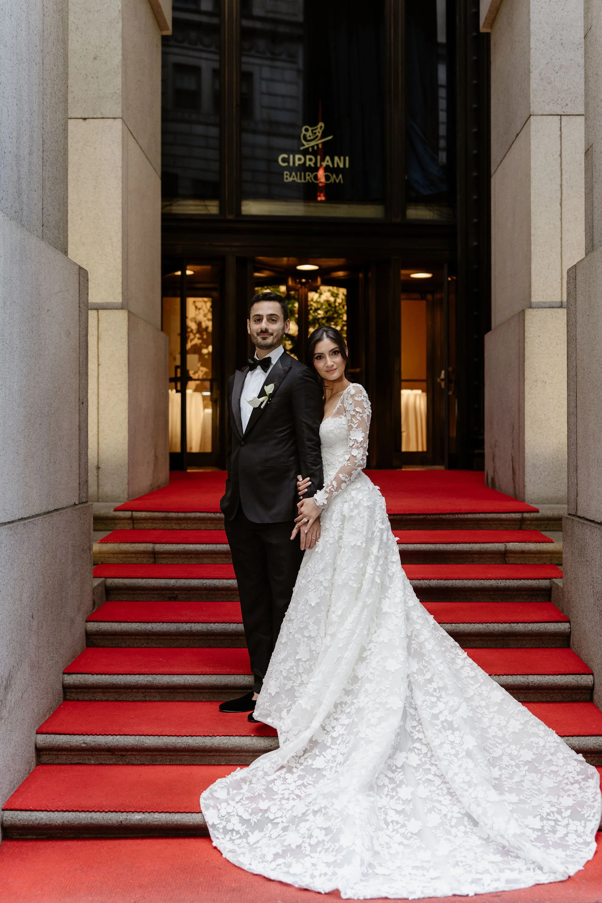 A bride and groom standing on a red carpet outside a building with a sign that reads Cipriani Ballroom. The bride is wearing a white lace wedding gown and the groom is dressed in a black tuxedo with a white shirt and black bow tie. They are posing together, looking at the camera.