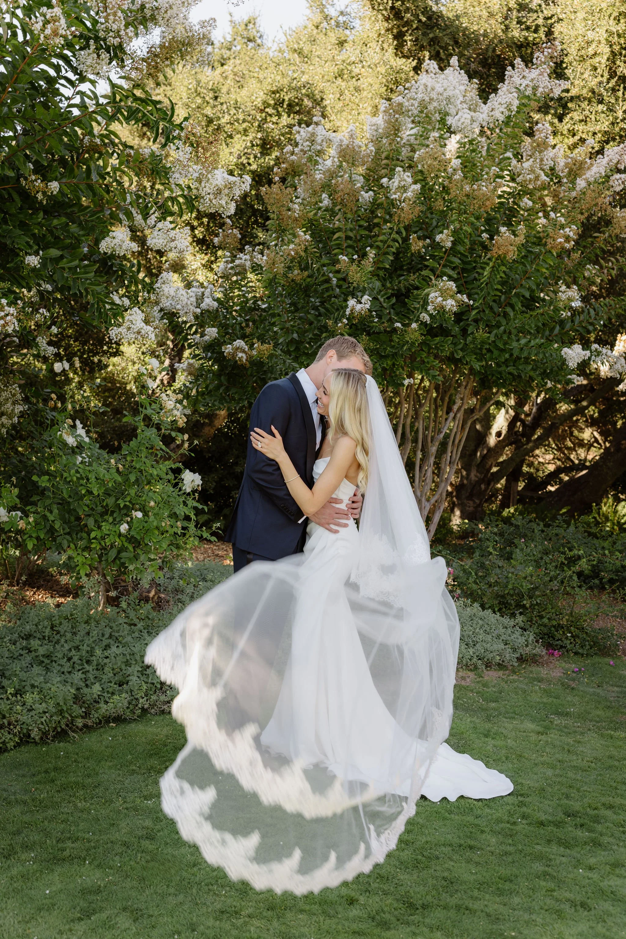 A bride and groom share a kiss outdoors beneath a flowering tree, with the bride in a white wedding dress and veil, and the groom in a dark suit, during a wedding celebration.