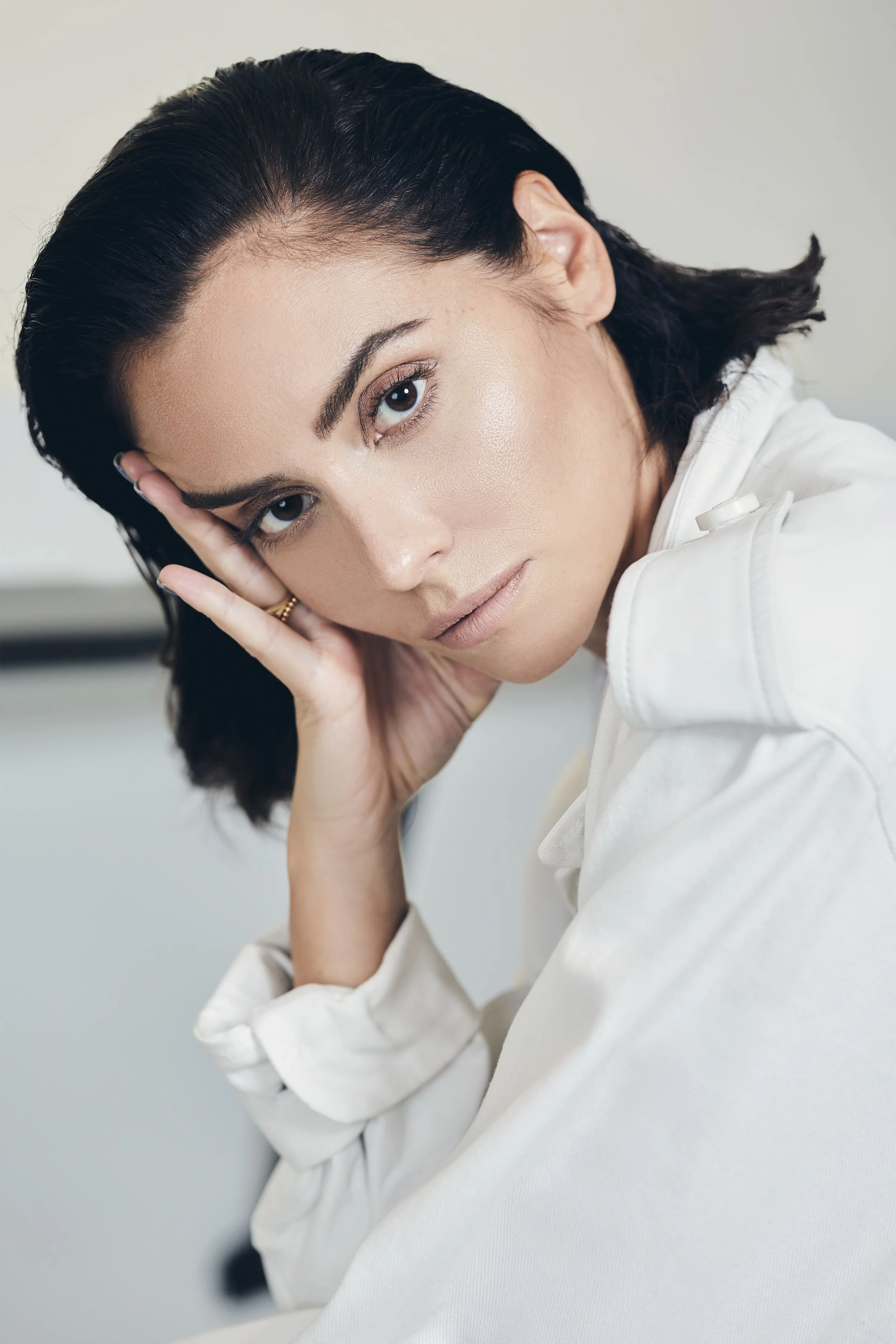 A woman with dark hair and light makeup, resting her head on her hand, looking at the camera with a neutral expression, wearing a light-colored jacket.