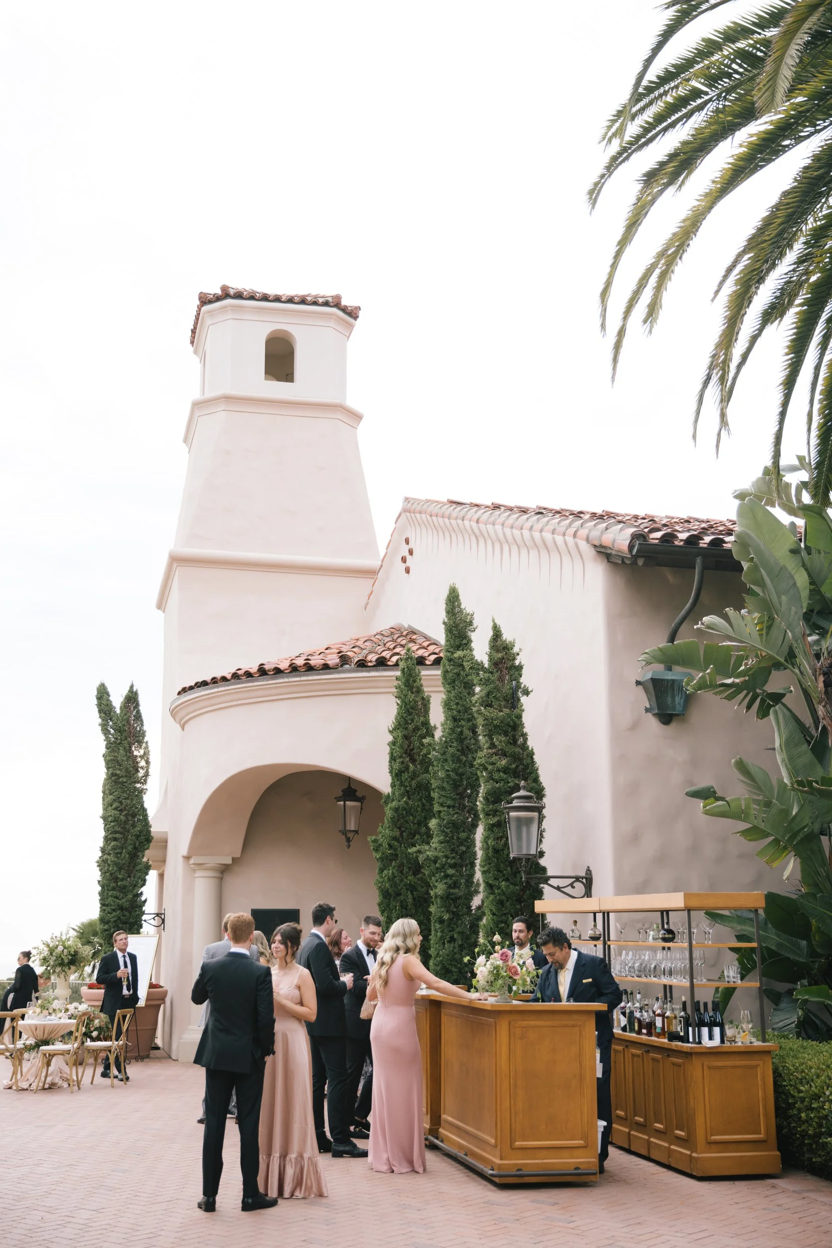 People dressed in formal attire socializing at an outdoor event under a building with a tall white tower and red tile roof, with greenery including palm trees and other plants nearby.