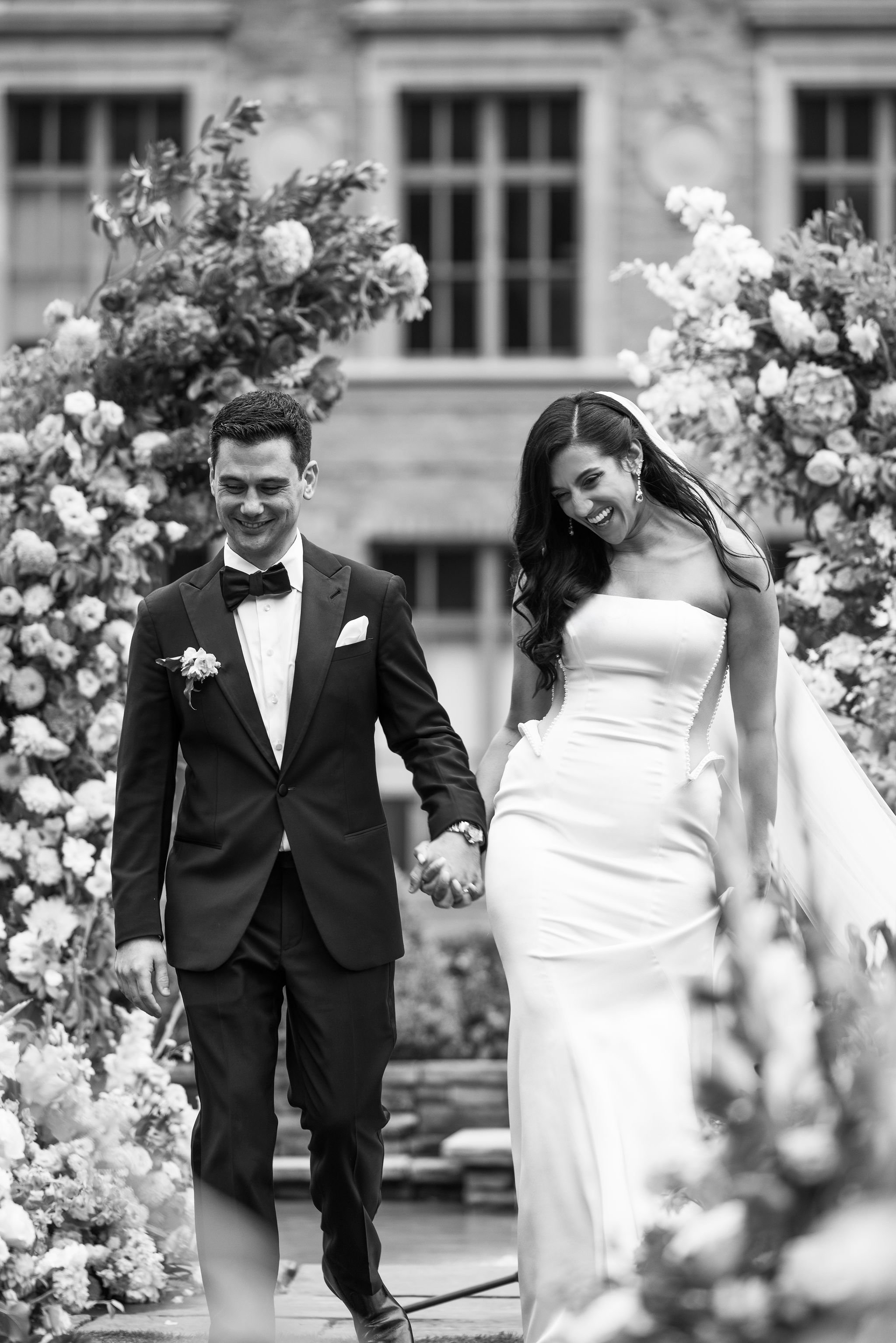 Black and white photo of a happy bride and groom walking hand in hand, surrounded by floral decorations, outside a large building.