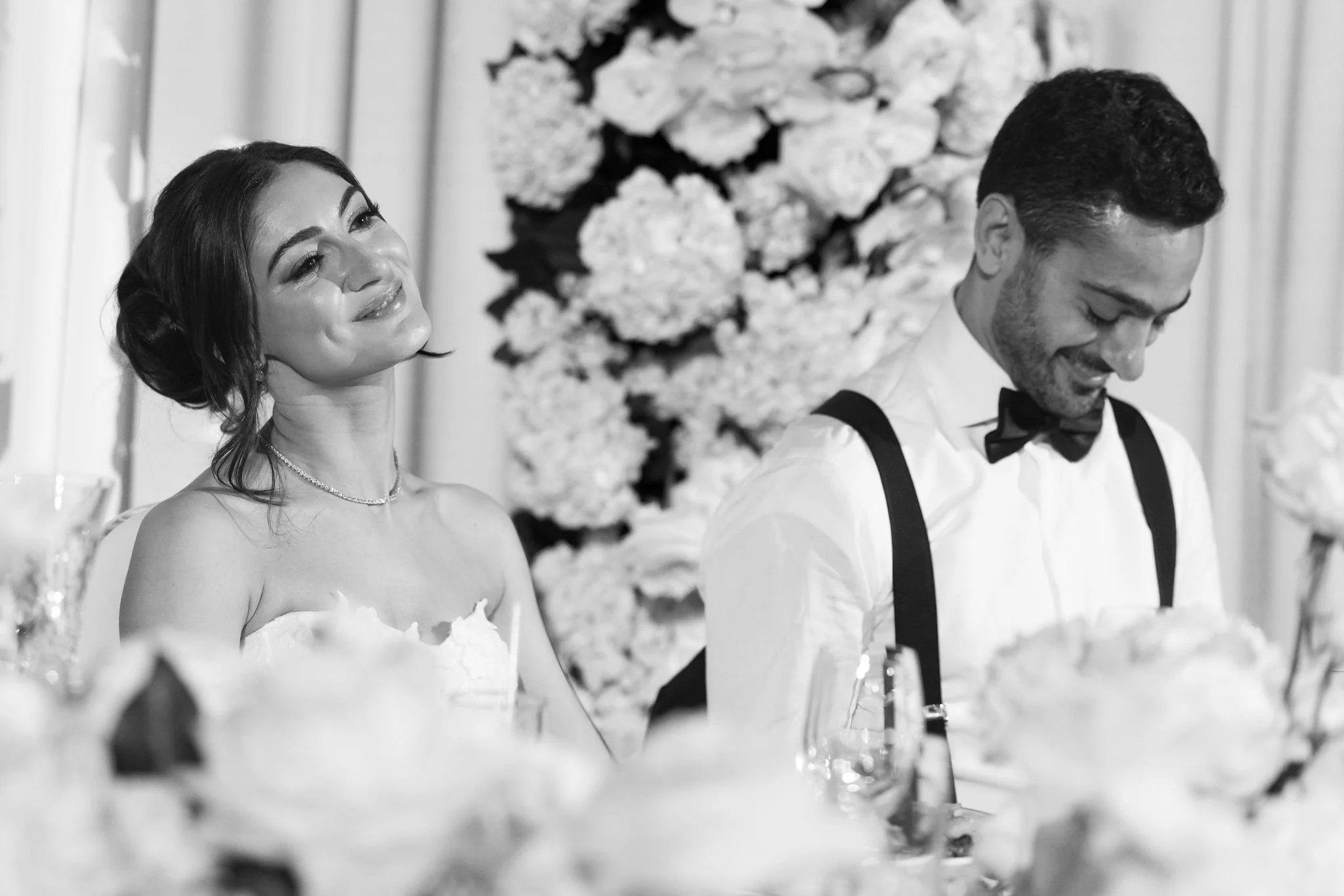 A black-and-white photo of a bride and groom at a wedding reception, sitting at a decorated table.