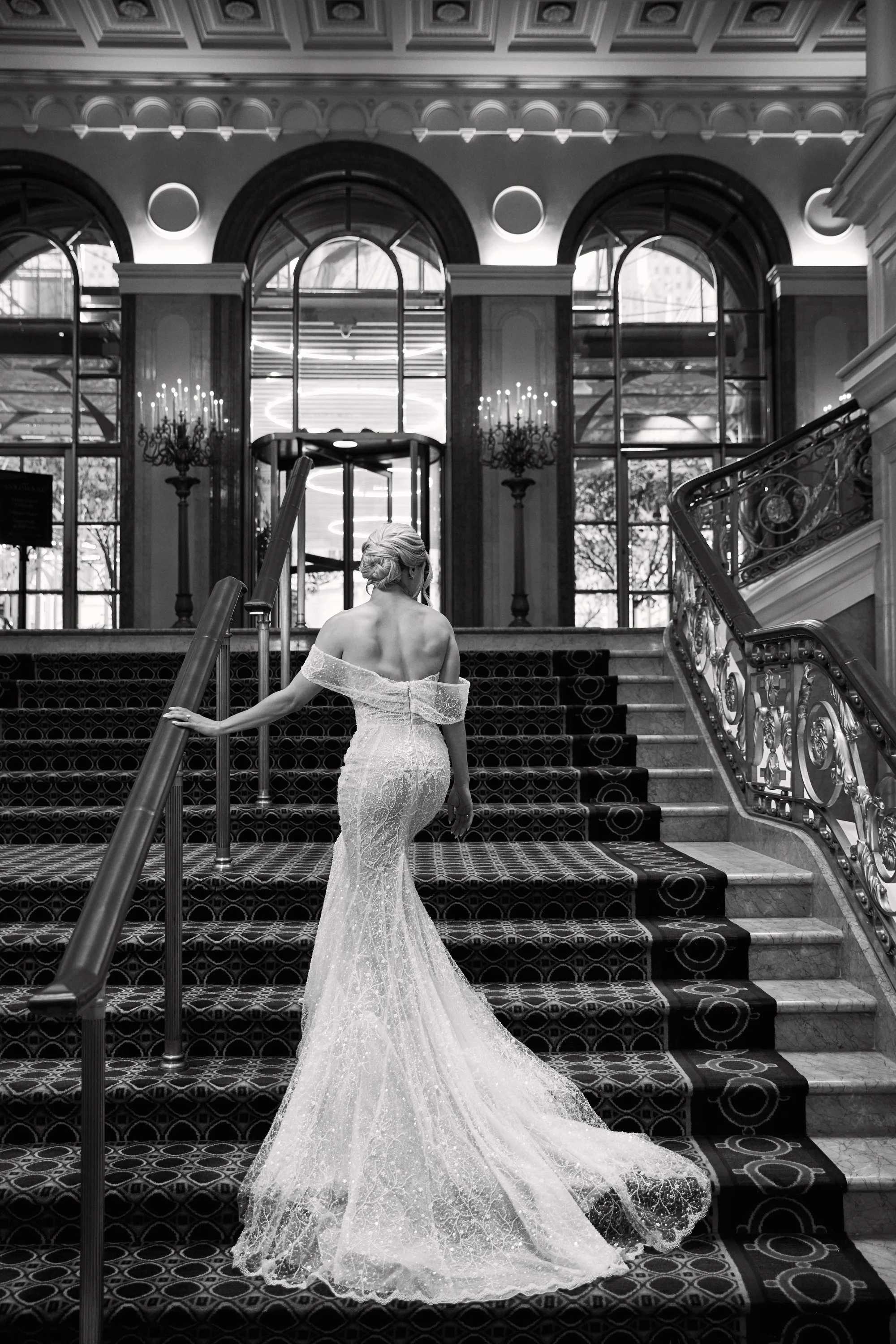 A woman in a strapless, form-fitting lace wedding gown with a long train, standing on a staircase inside an ornate building with large arched windows, marble stairs, and elegant chandeliers.