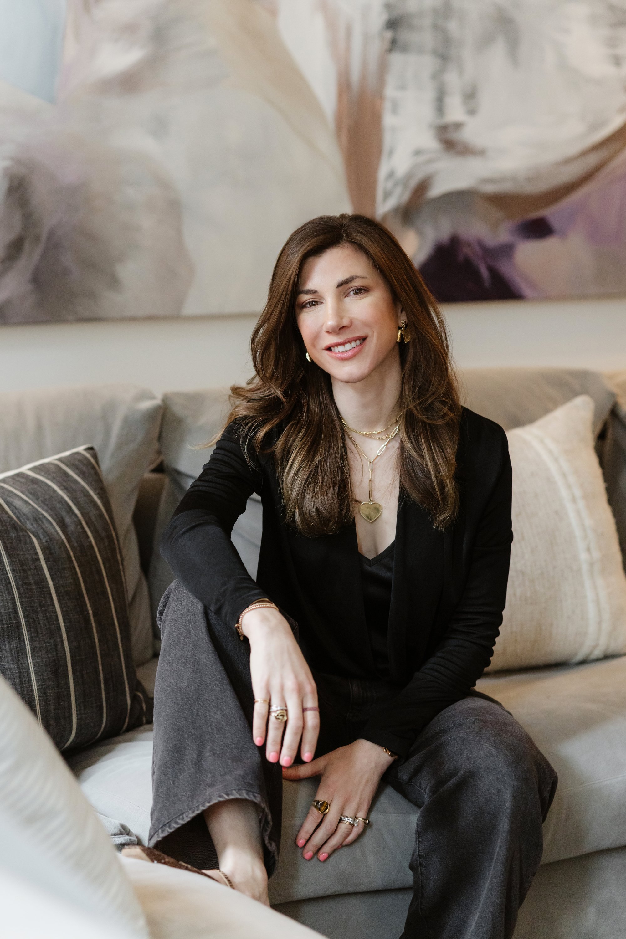 A woman with shoulder-length brown hair, wearing a black blazer and gray pants, sitting on a sofa with patterned pillows, smiling at the camera in a cozy living room with a large abstract painting in the background.