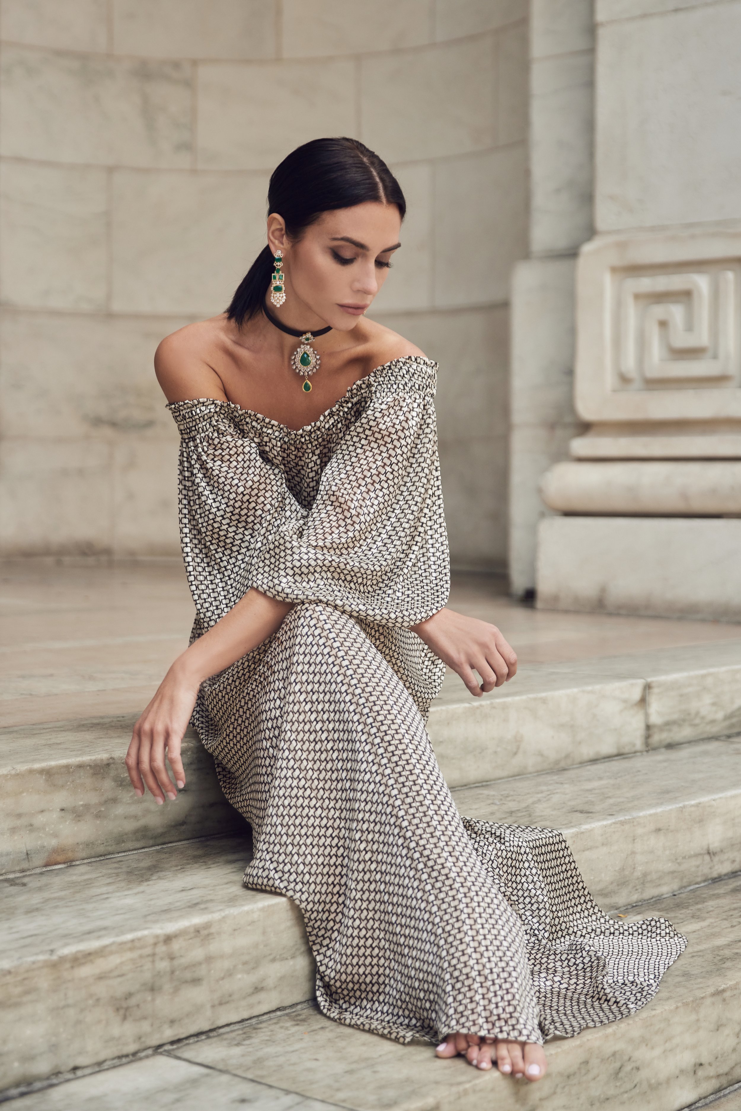 A woman with dark hair styled in a sleek bob, sitting on stone steps inside a building with classical architecture. She is wearing a patterned off-the-shoulder dress and elegant jewelry, including a black choker with a large green pendant, matching e