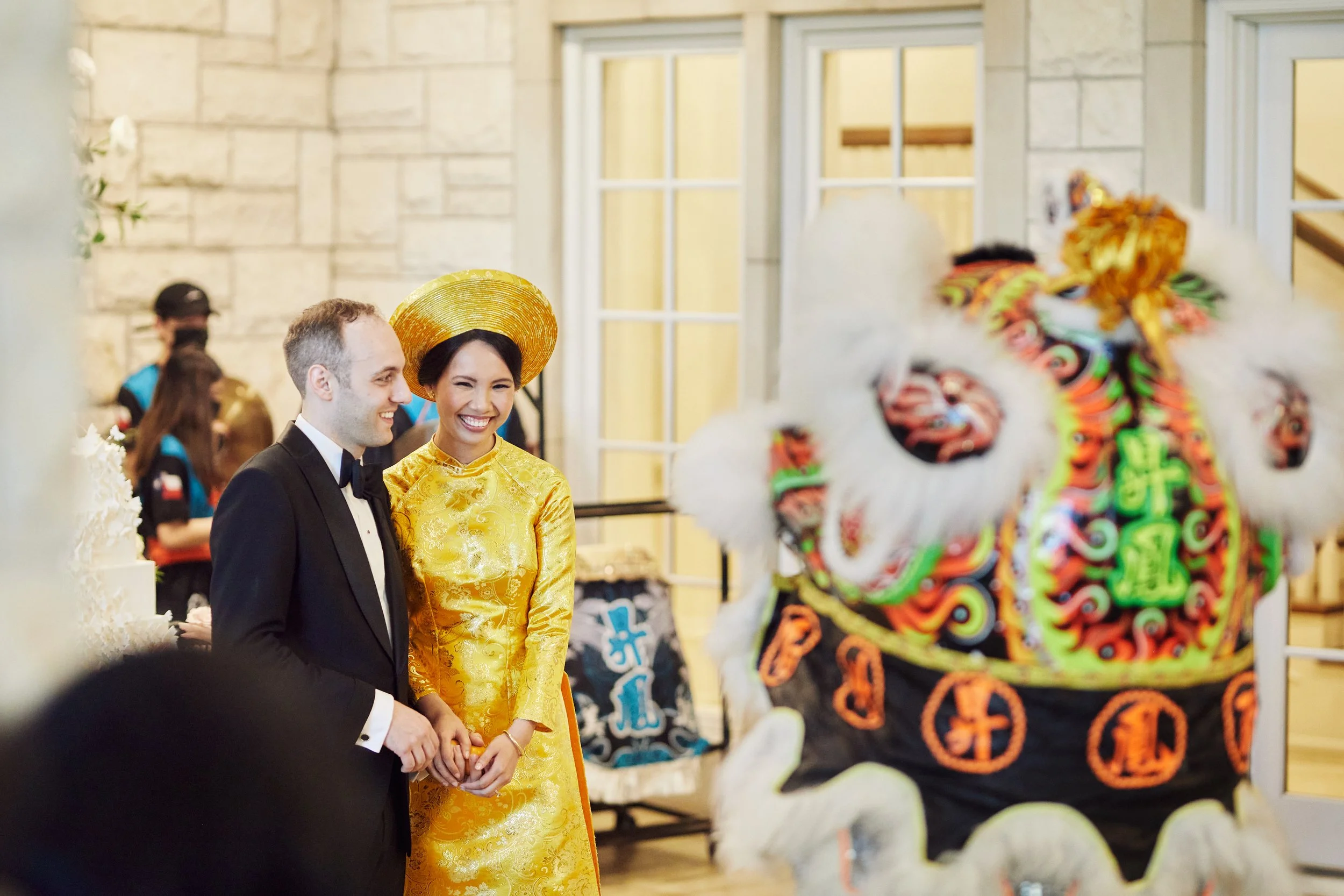 A bride and groom are smiling and holding hands during their wedding reception, with a lion dance costume in the foreground and decorations in the background.
