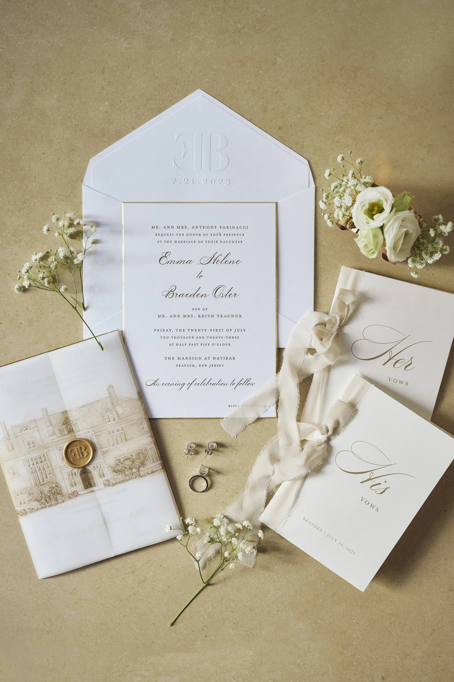 Wedding invitation suite, including an envelope with embossed monogram and date, a printed invitation with gold accents, a small bouquet of white flowers, a sealing wax seal with initials, and a pair of earrings.
