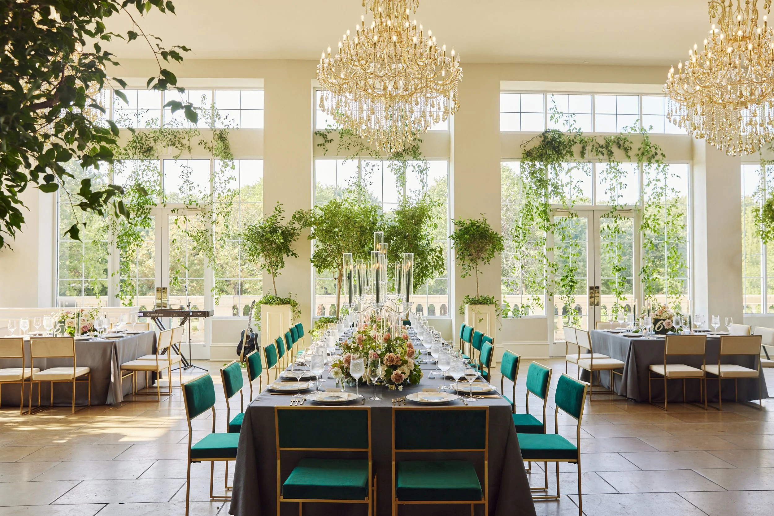 Elegant event space with high ceilings, large windows, and two ornate chandeliers. A long table with teal chairs is decorated with floral centerpieces, wine glasses, and tableware.