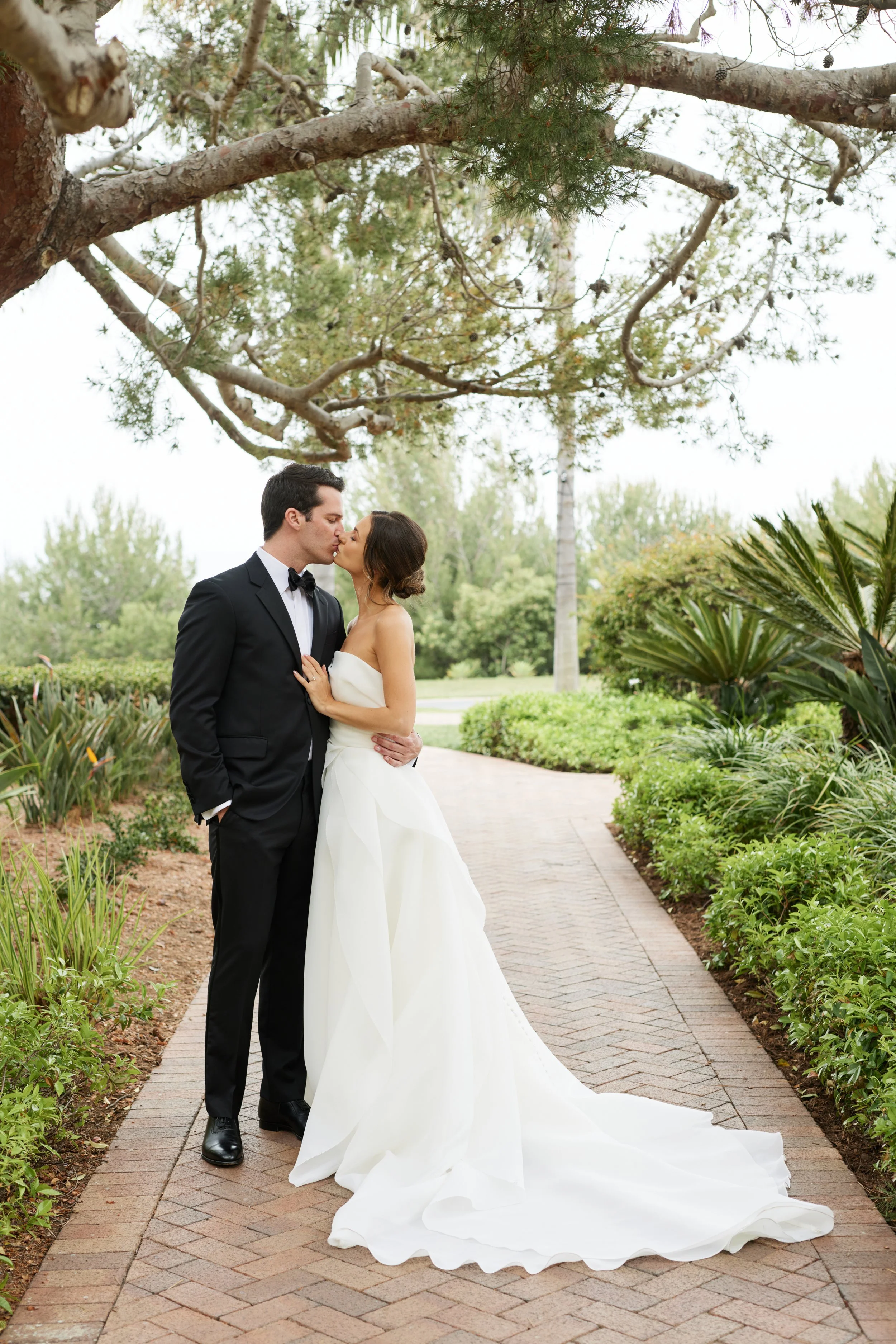 A bride and groom share a kiss in a garden pathway surrounded by green plants and trees. The bride wears a strapless white wedding gown with a train, and the groom wears a black tuxedo with a bow tie.