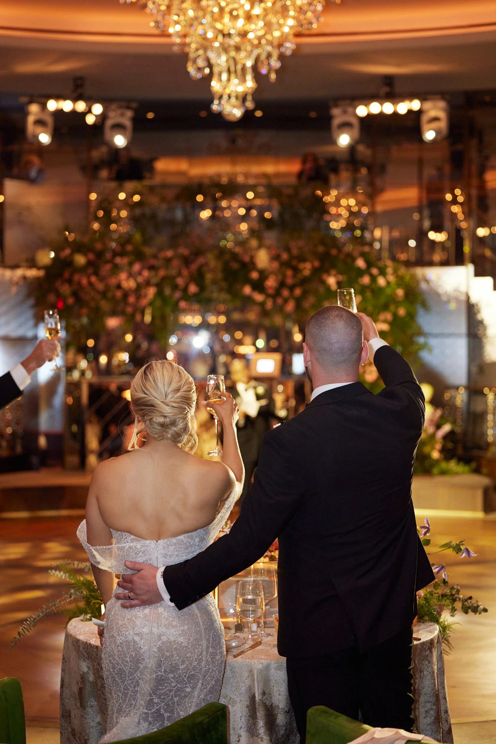 A bride and groom celebrating at their wedding reception with raised glasses, standing near a decorated table, with a chandelier and floral arrangements in a warmly lit venue.
