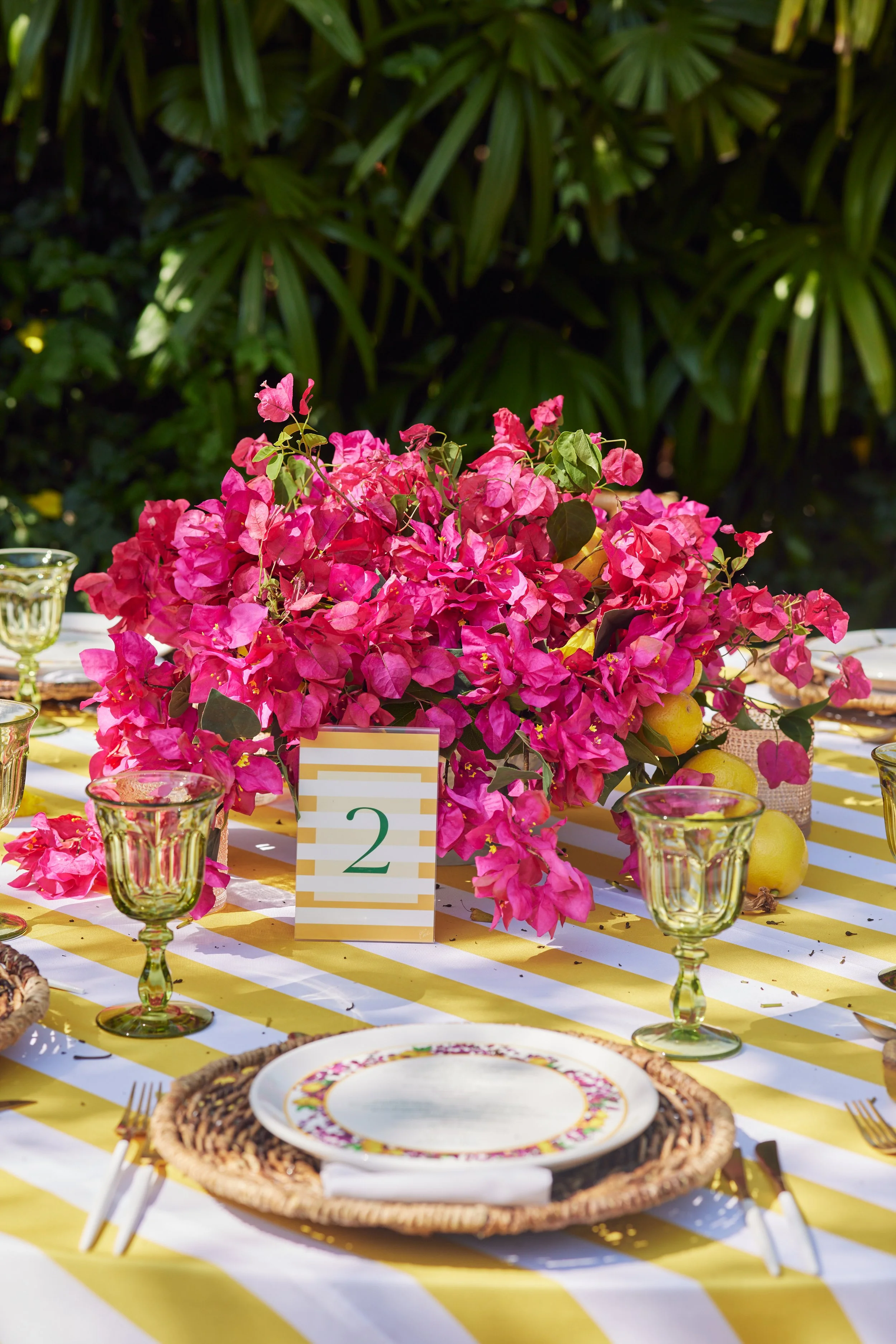 A brightly decorated outdoor table with a centerpiece of pink flowers, surrounded by glasses, plates, and utensils, with a yellow and white striped tablecloth. In the background, green foliage is visible.