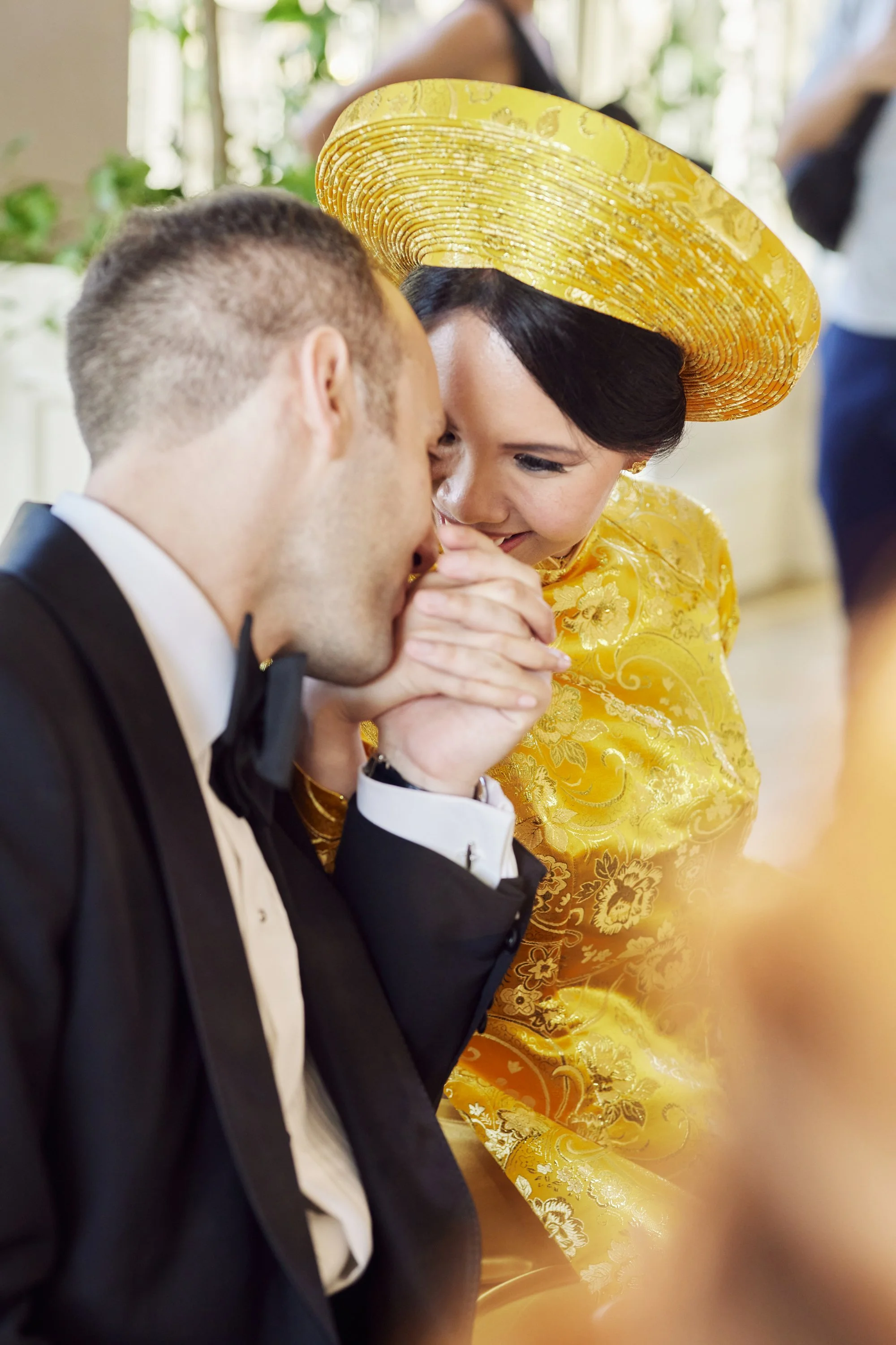 A woman dressed in a yellow traditional Mexican dress with a large yellow sombrero, holding hands and touching foreheads with a man in a black tuxedo, during a celebration or wedding.
