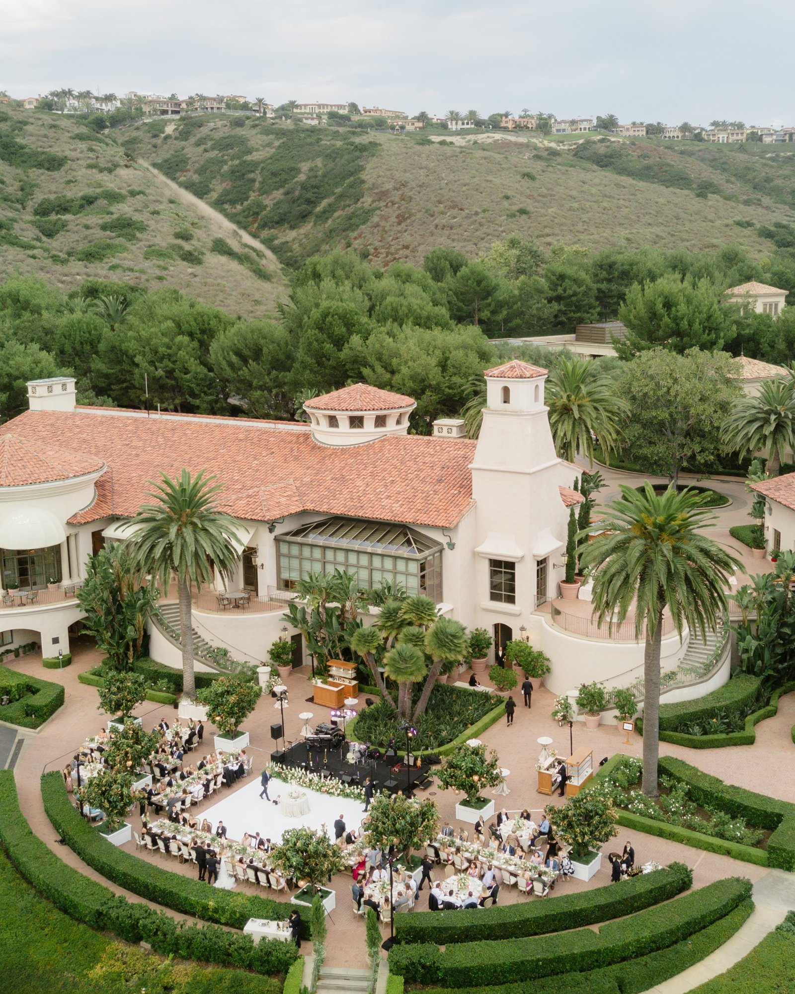 A large outdoor wedding reception taking place at a lush, green estate with a white building featuring a red-tiled roof. Guests are seated around round and rectangular tables, and there is a grand piano on a dance floor. The landscape includes well-manicured hedges, tall palm trees, and hills with houses in the background under a cloudy sky.