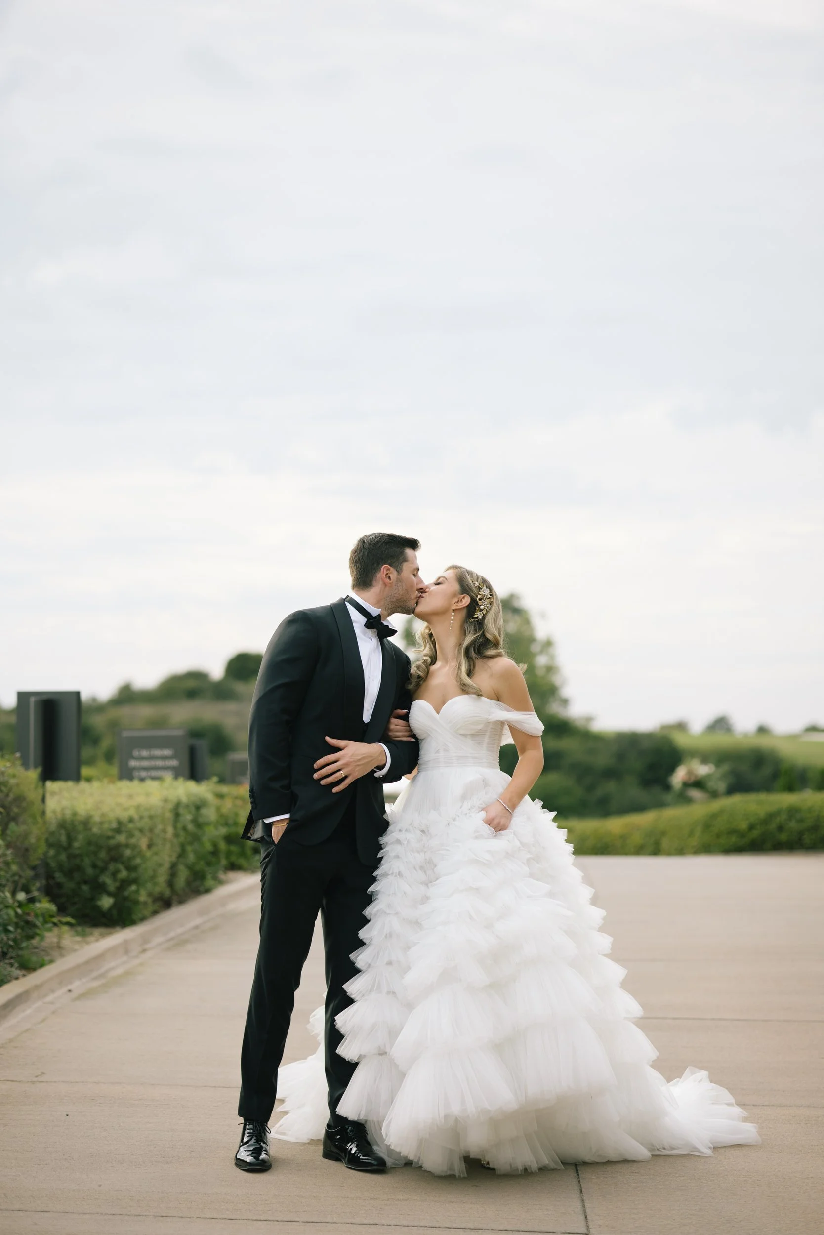 A newlywed couple sharing a kiss outdoors on a cloudy day. The groom wears a black tuxedo with a bow tie, and the bride wears a white strapless wedding gown with ruffled layers. They are standing on a paved path with greenery and trees in the background.