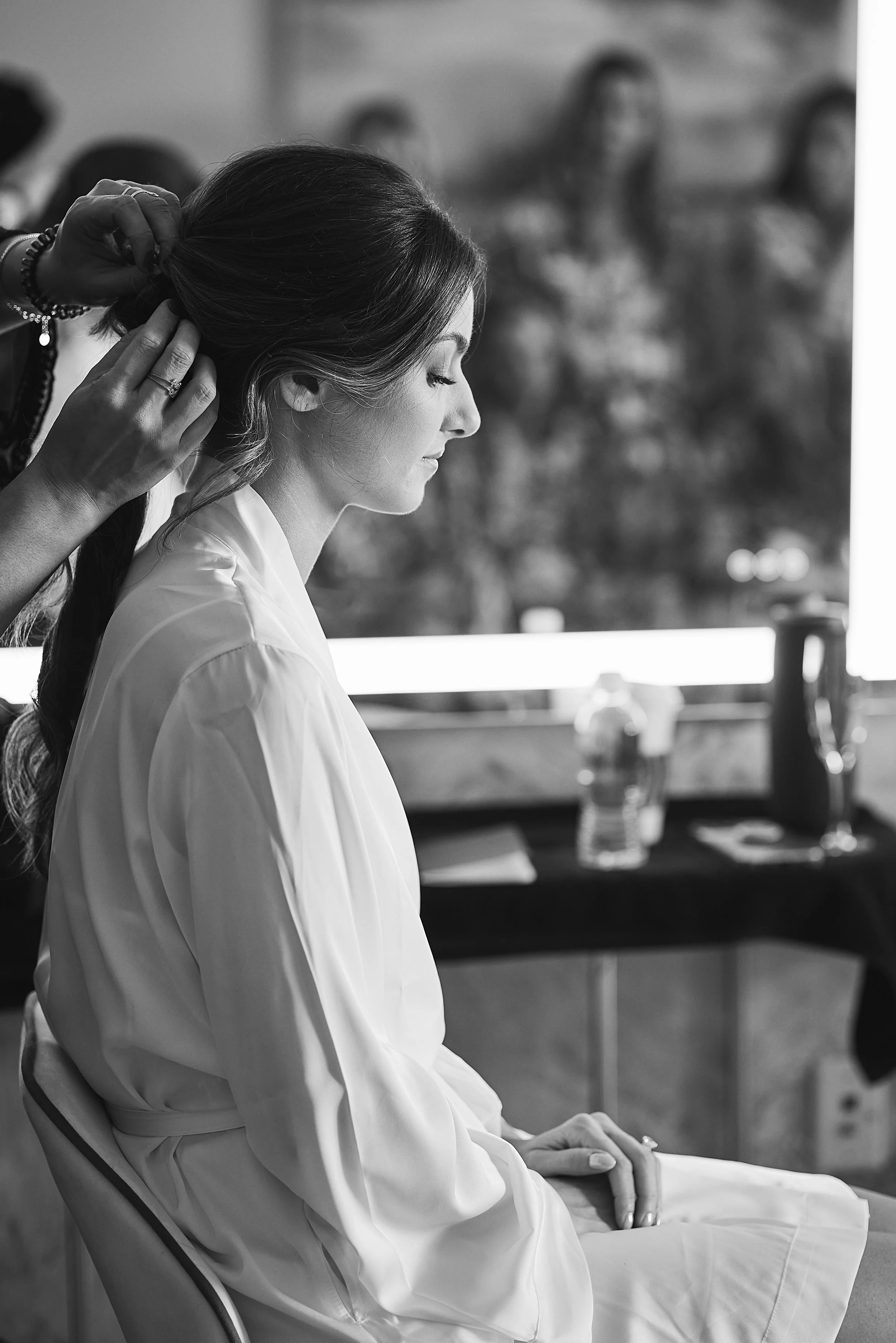 A woman dressed in a white outfit, seated with her eyes closed, having her hair styled by someone in the background. Each person appears to be in a relaxed setting.