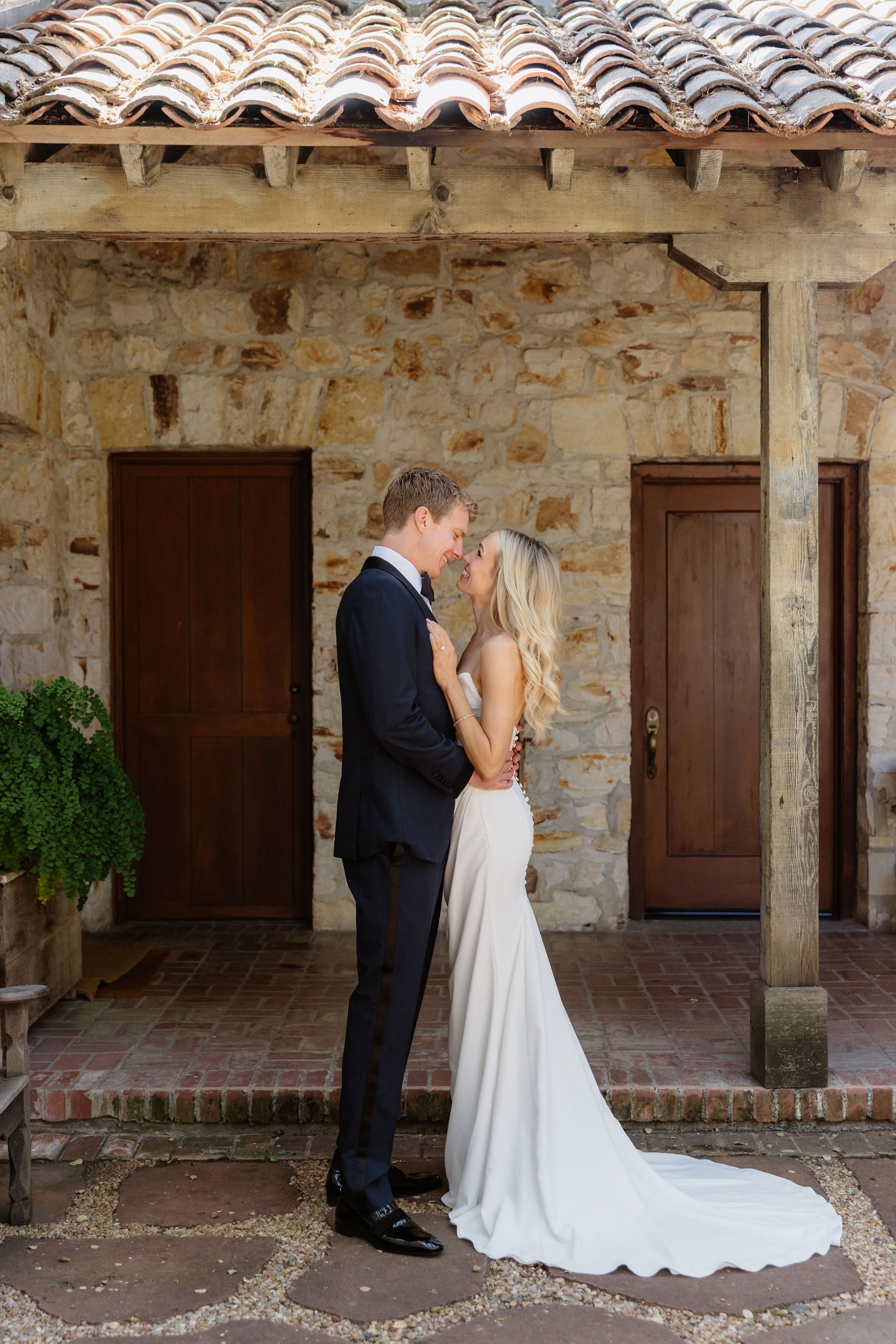 A bride and groom dance closely in front of a rustic stone wall with wooden doors, under a wooden beam with a tiled roof, in an outdoor setting.